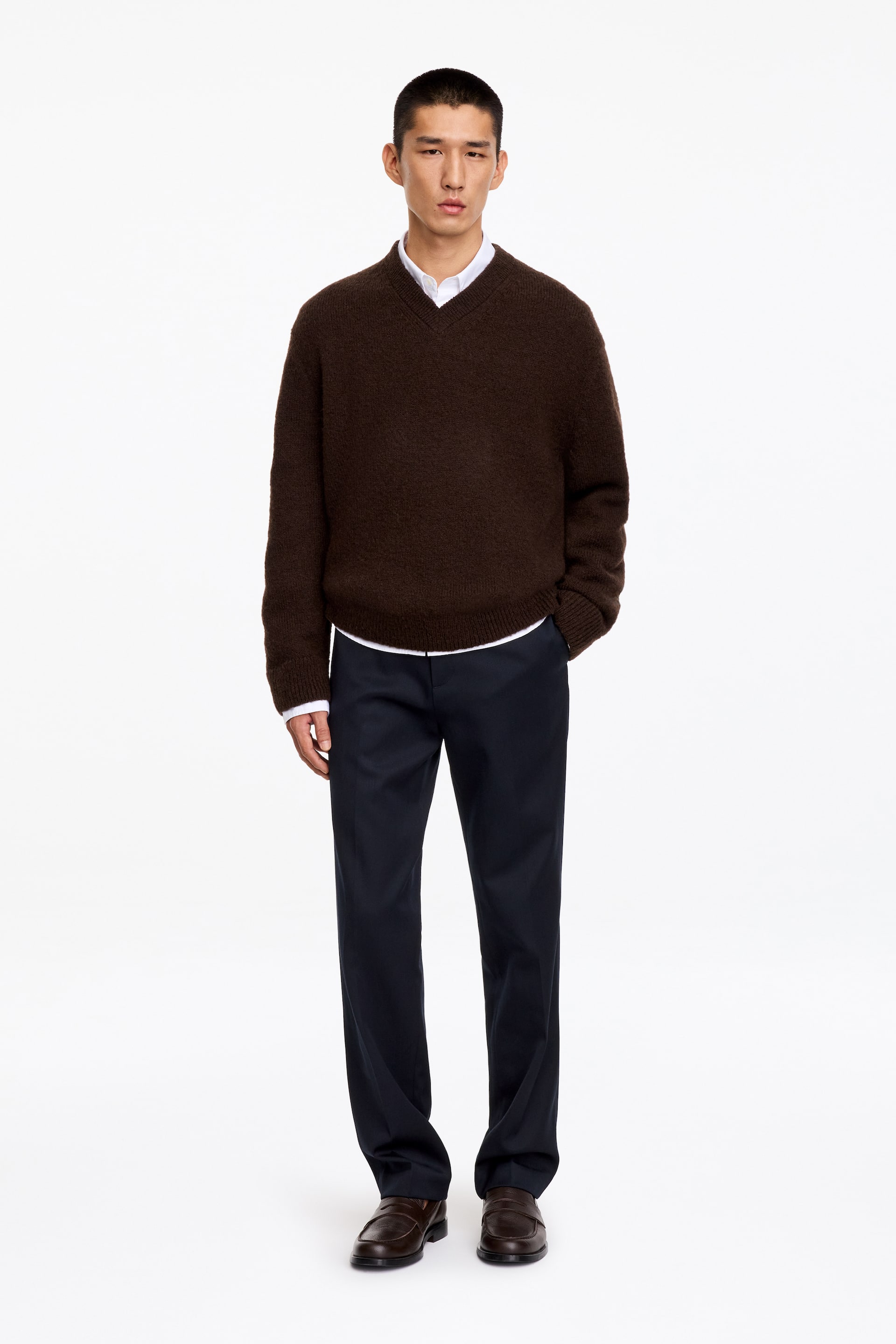 Agrandir l'image: Le mannequin porte un pantalon en laine et coton d’une teinte bleu marine foncé, doté d’une coupe droite et de plis marqués subtils. Le pantalon est associé à un pull col V marron foncé, une chemise blanche à col et des mocassins en cuir marron.