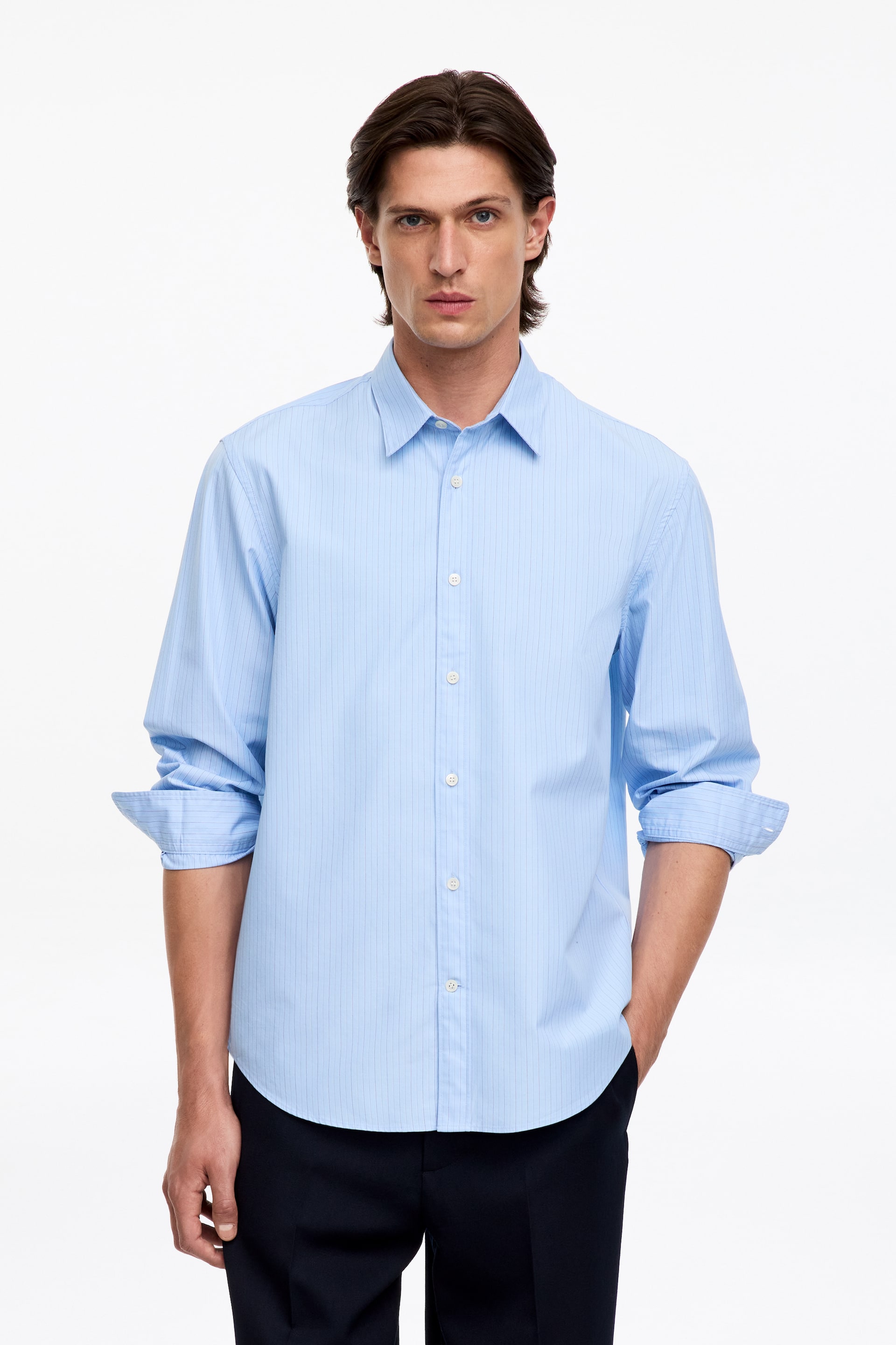 Agrandir l'image: Le mannequin porte une chemise boutonnée bleu clair à fines rayures verticales et à col classique. La chemise présente des boutons blancs sur le devant, et ses manches sont retroussées jusqu’aux avant-bras.