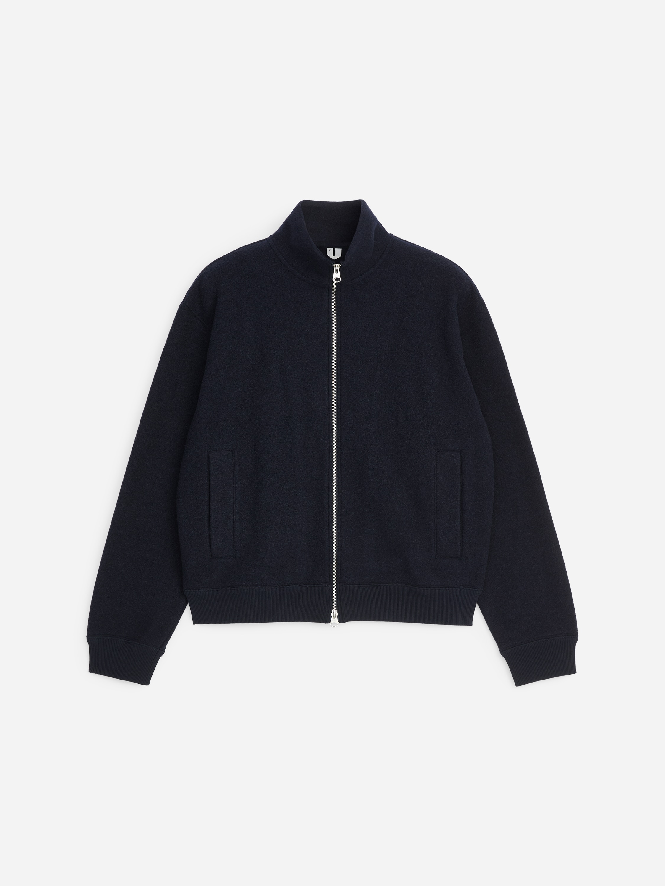 La modelo lleva puesta una chaqueta de mezcla de lana en azul marino oscuro, con cuello alto, cierre de cremallera bidireccional plateada y acabados acanalados. La chaqueta se combina sobre una camiseta blanca y se lleva con pantalones gris oscuro.