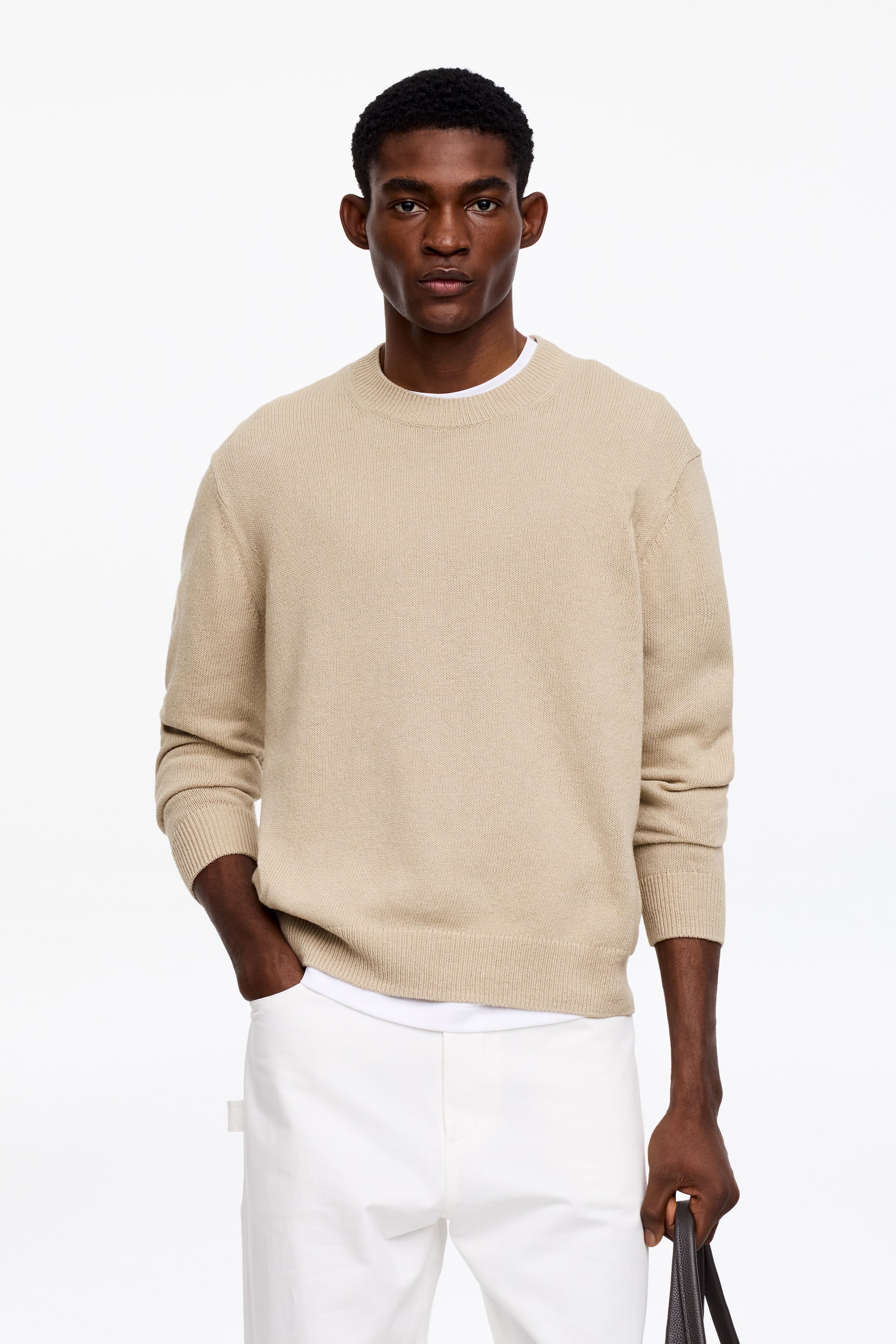 Grotere afbeelding bekijken: Een beige trui met ronde hals wordt aan de voorkant gepresenteerd, met een gebreide textuur, geribde boorden en een geribde zoom, en wordt over een wit T-shirt gedragen.