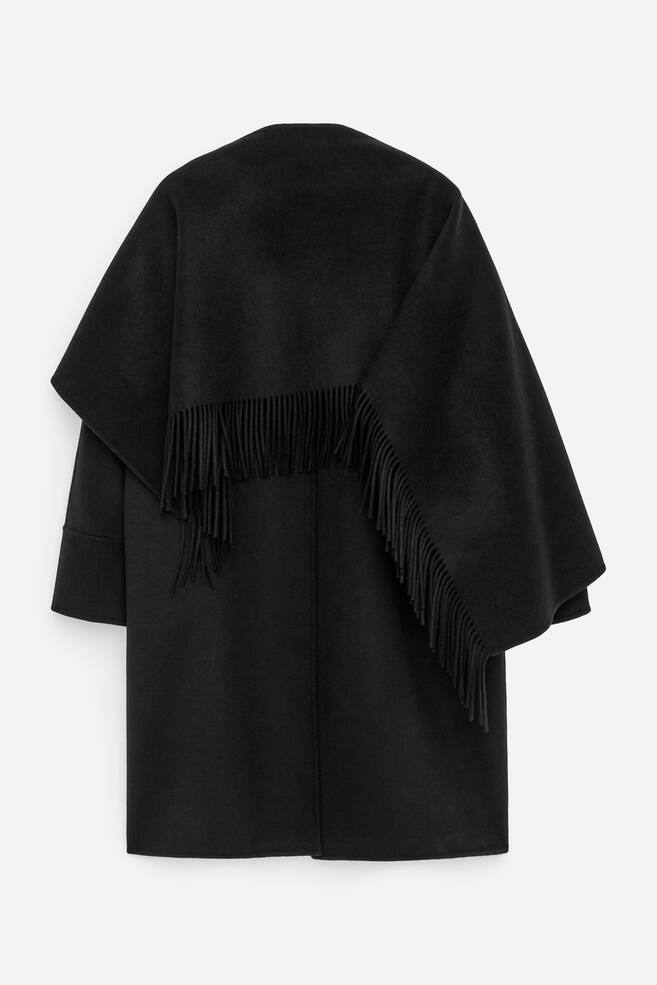 En svart cape-kappa visas bakifrån och framhäver ett slätt, mörkt material. En fransad scarf är draperad över axlarna och övre delen av ryggen, med fransarna synligt hängande ner. - 1