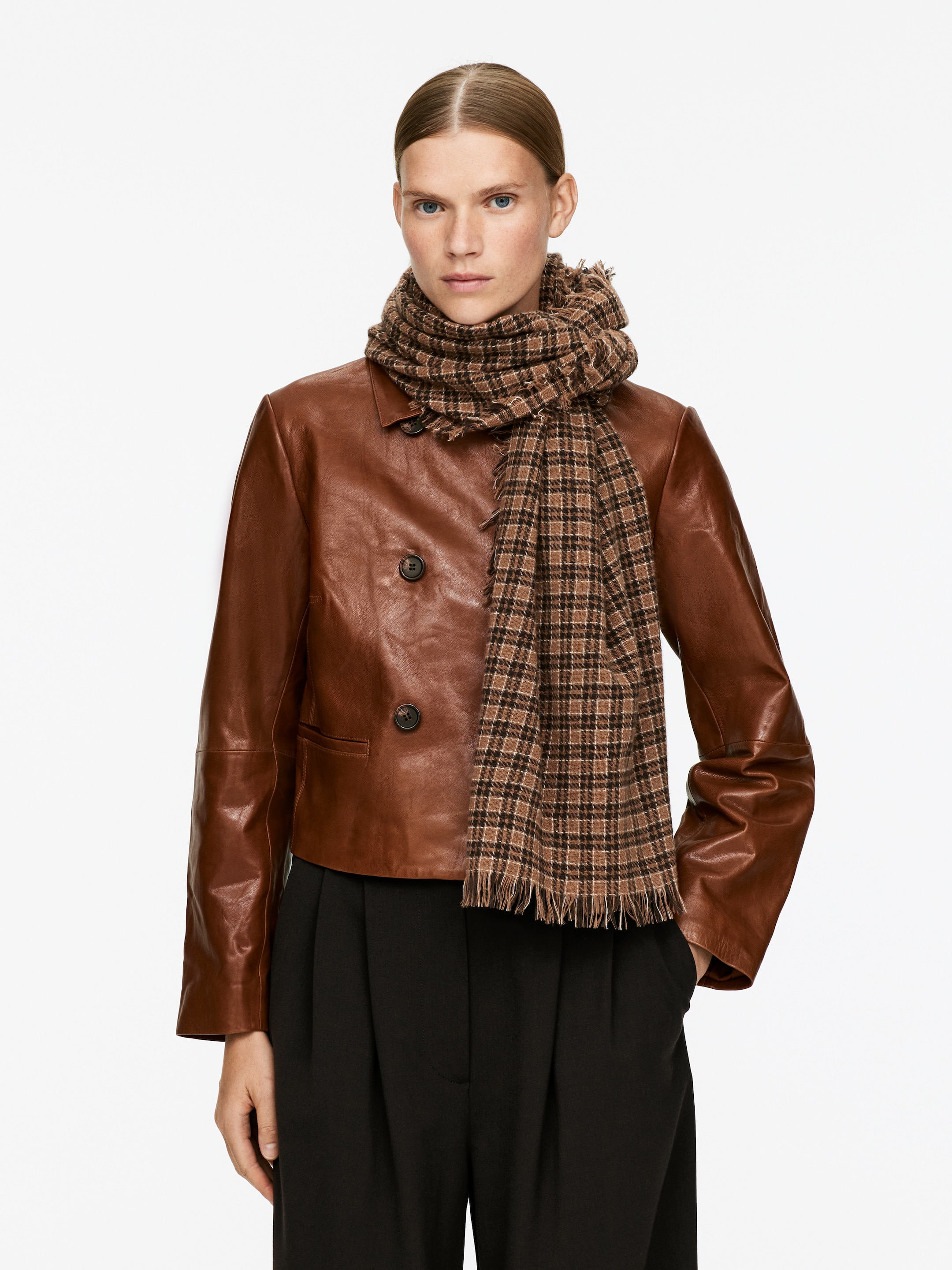 View larger image: A brown leather cropped double-breasted jacket is presented from the front, featuring two dark buttons and a pointed collar. A brown and black plaid scarf with frayed edges is draped around the neck over the jacket, with black wide-leg pants visible below.
