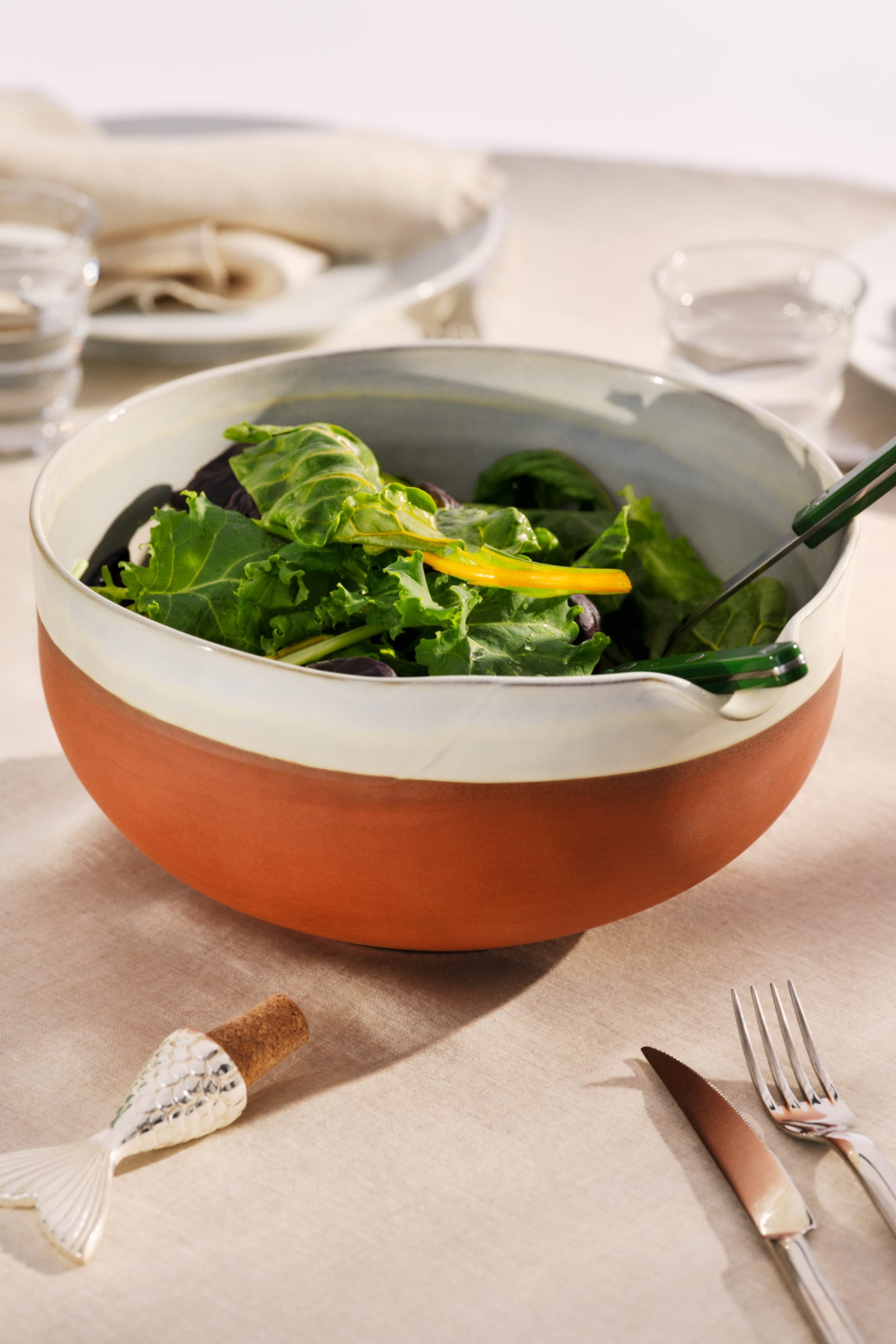 Ver imagen más grande: Un cuenco de ensalada redondo de dos tonos presenta una sección inferior de terracota y un borde superior blanco roto. Contiene una ensalada verde mixta con tiras de pimiento amarillo, y unas pinzas verdes para servir descansan en dos muescas integradas en el borde.