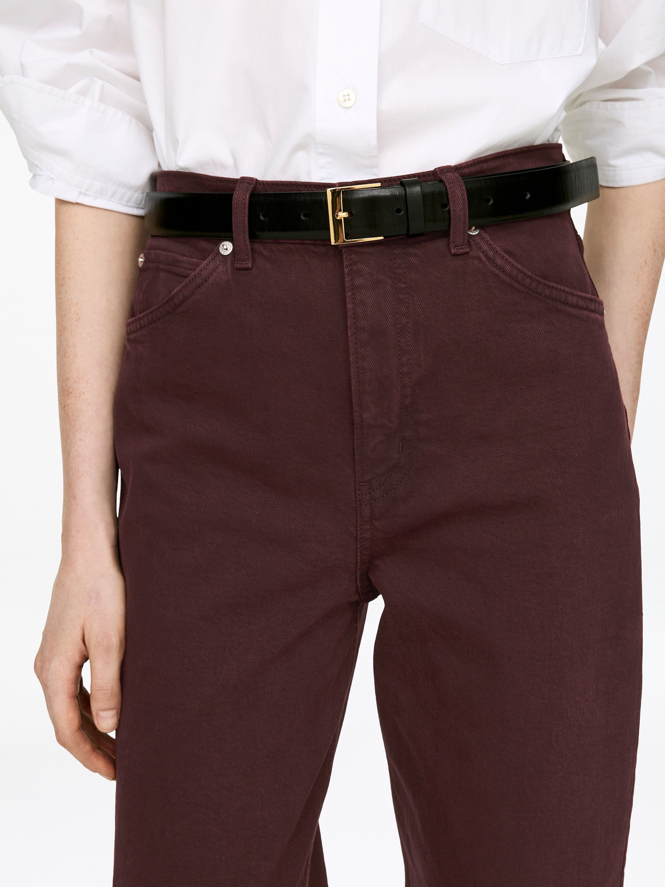 Visualizza immagine più grande: Pantaloni LUPINE rosso-bruni scuri sono indossati dalla modella, caratterizzati da una vita alta, tasche anteriori con rivetti metallici e passanti per cintura, con una cintura nera e fibbia dorata infilata attraverso di essi. Una camicia bianca è infilata nei pantaloni.