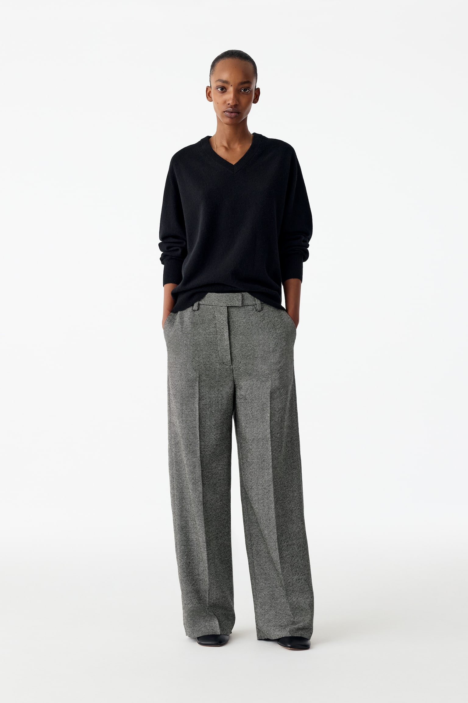 De grijze broek met textuur is van voren gefotografeerd en toont een rechte pijp met scherpe geperste vouwen die in het midden van elk been lopen. Ze zijn voorzien van een gestructureerde tailleband met riemlussen en subtiele steekzakken aan de zijkant. - 1
