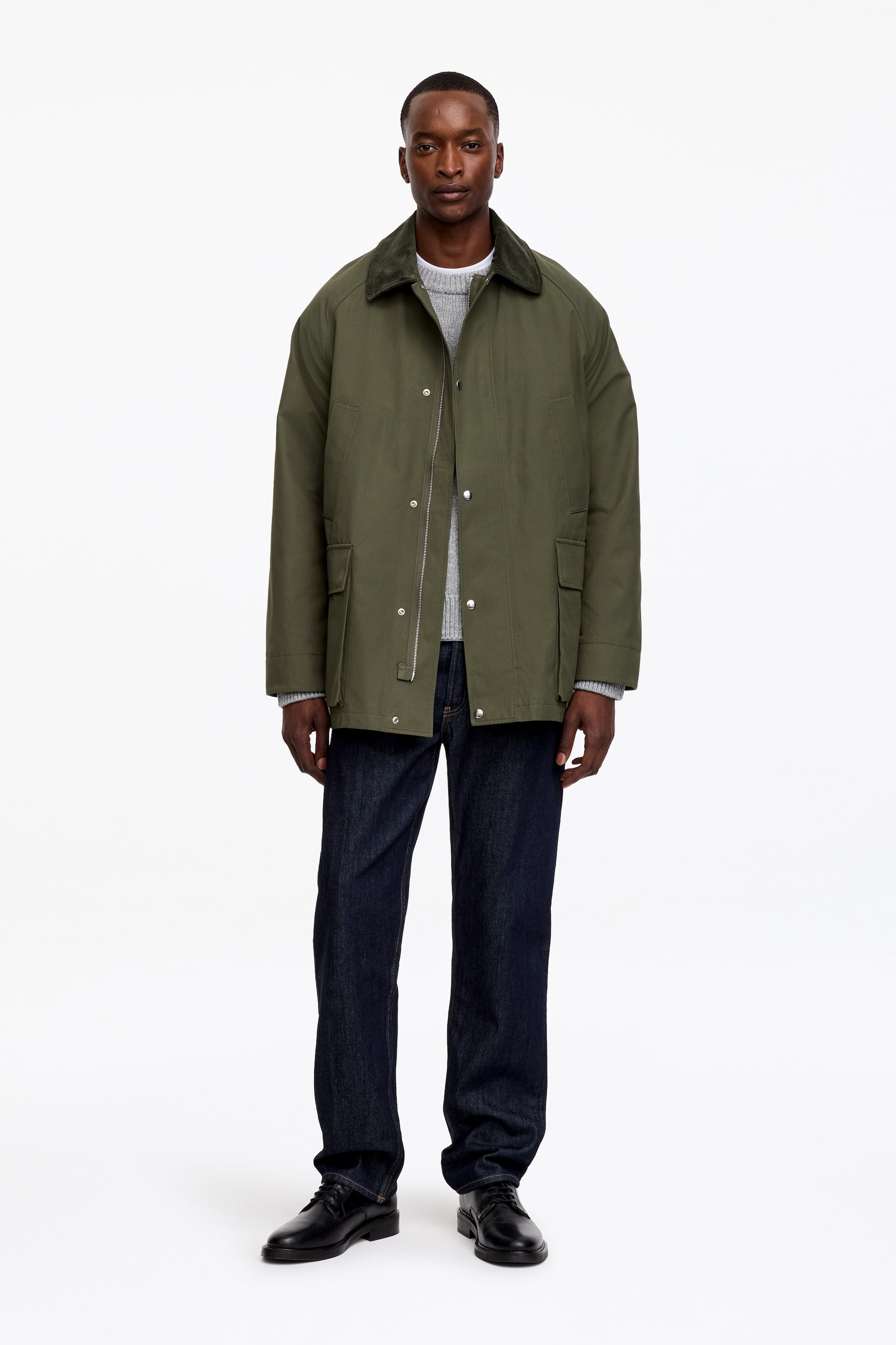 View larger image: An olive green mid-length coat with a waxed finish is being worn by the model over a light grey jumper. The coat features a dark corduroy collar, raglan-cut sleeves, and a front placket with popper buttons that partially covers a two-way zip. Two slanted welt pockets and two patch pockets with flaps and popper button closures detail the front of the coat.