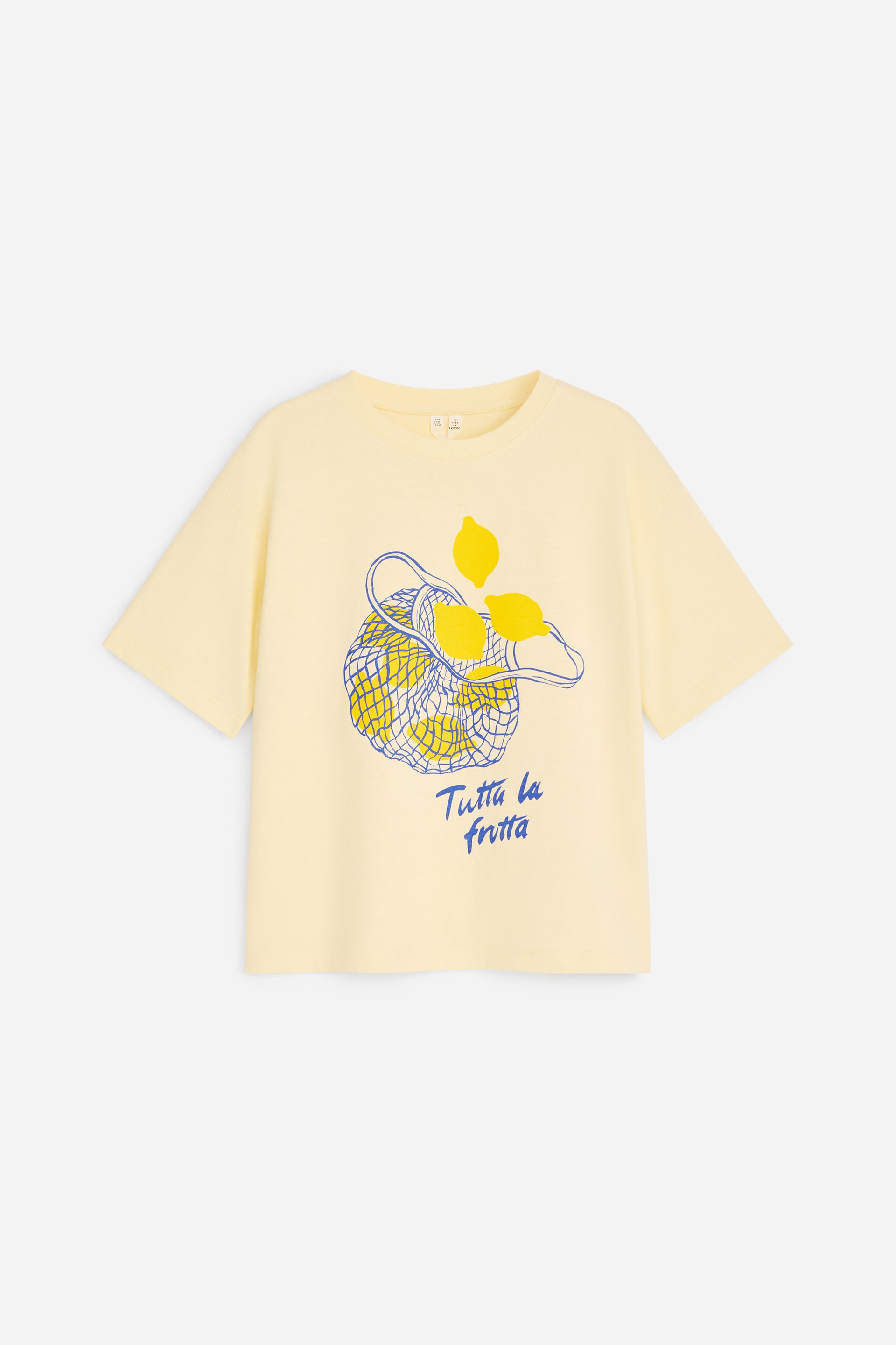 Grotere afbeelding bekijken: Een lichtgele t-shirt wordt van voren getoond, met een oversized pasvorm, licht afhangende schouders en korte mouwen. Het heeft een klassieke ronde hals en een centrale afbeelding van gele citroenen in een blauw netzakje, met daaronder de tekst 'Tutta la frutta' in blauw.