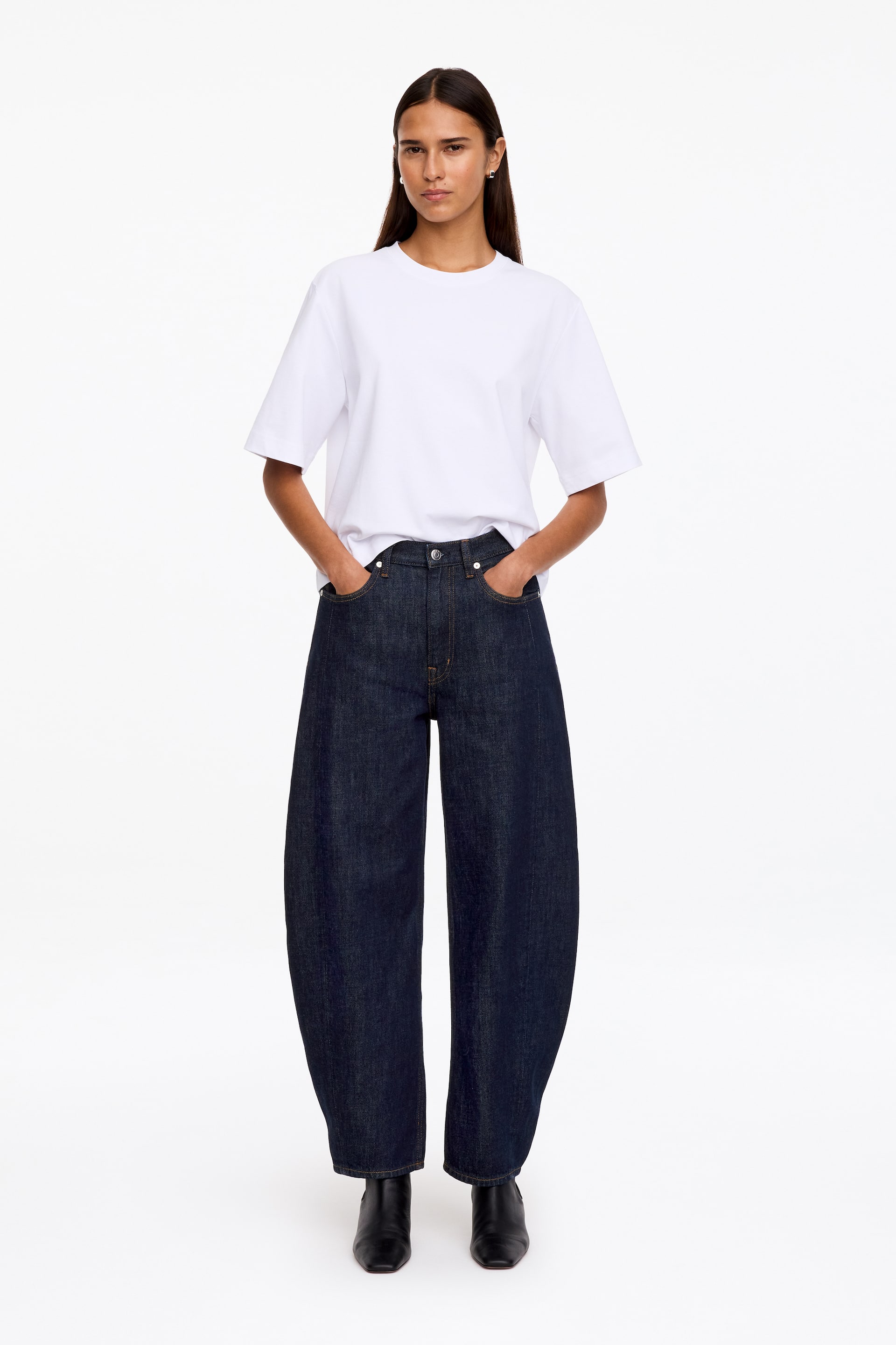 Ver imagen más grande: La modelo lleva unos vaqueros denim de color índigo oscuro con cintura de tiro medio y un corte relajado, con perneras recortadas y de silueta estructurada. La modelo aparece de pie con las manos en los bolsillos delanteros, combinando los vaqueros con una camiseta blanca metida por dentro y botas negras.