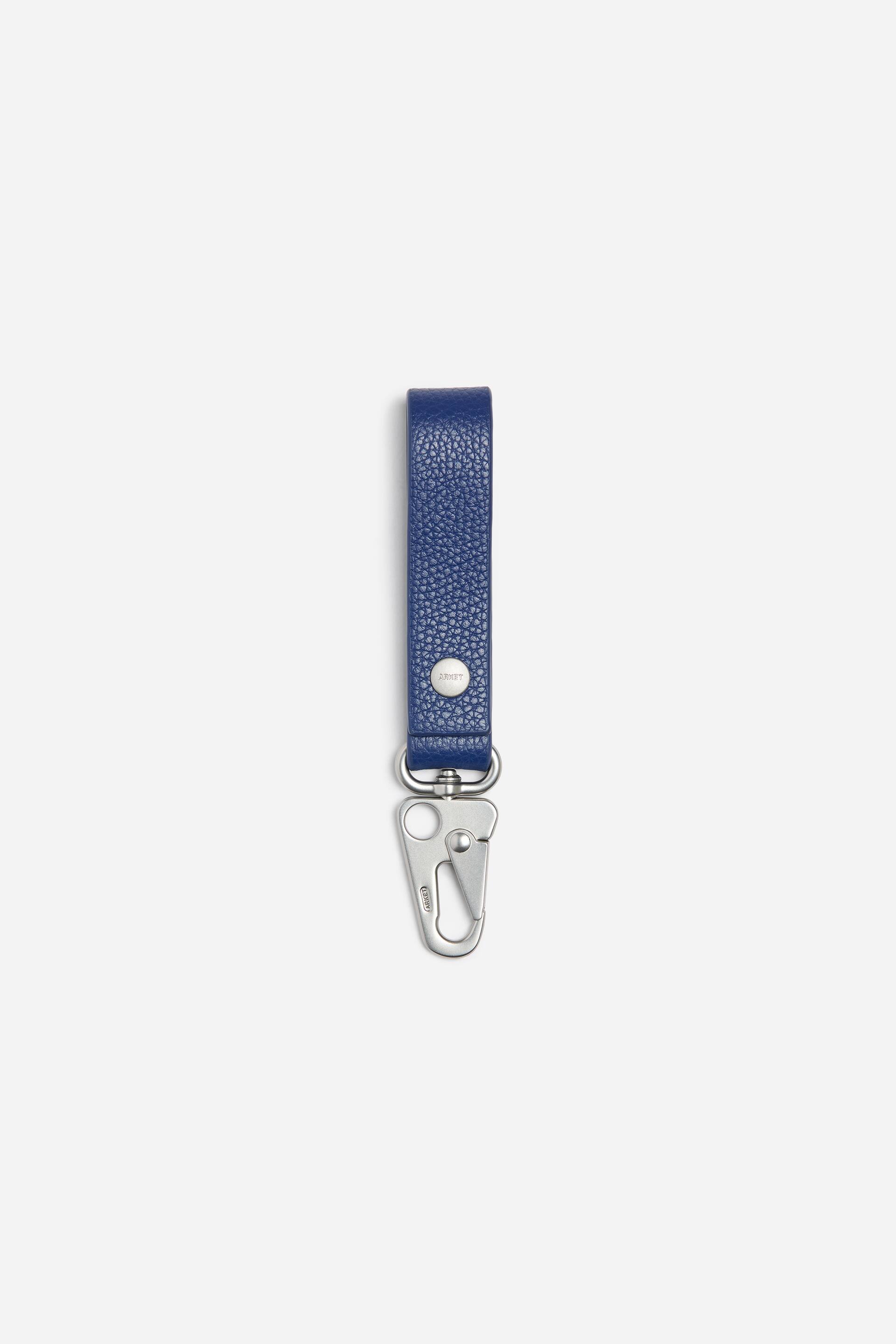 Grotere afbeelding bekijken: Een blauw, getextureerd leren sleutelhanger wordt van voren getoond, met een lusvormige band die is vastgezet met een zilverkleurige metalen drukknoop. De band is onderaan bevestigd aan een zilverkleurige metalen karabijnhaak met drukknoop.