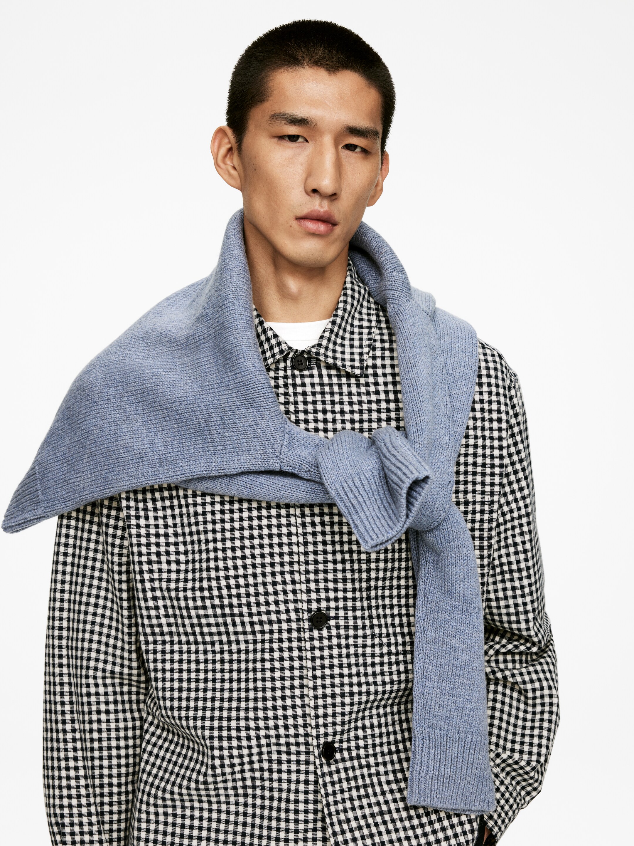 Visualizza immagine più grande: Una camicia a quadretti bianco e nero con bottoni, che presenta un colletto classico e bottoni neri, è indossata sotto un maglione azzurro lavorato a maglia, drappeggiato sulle spalle e annodato davanti, con una t-shirt bianca visibile sotto.