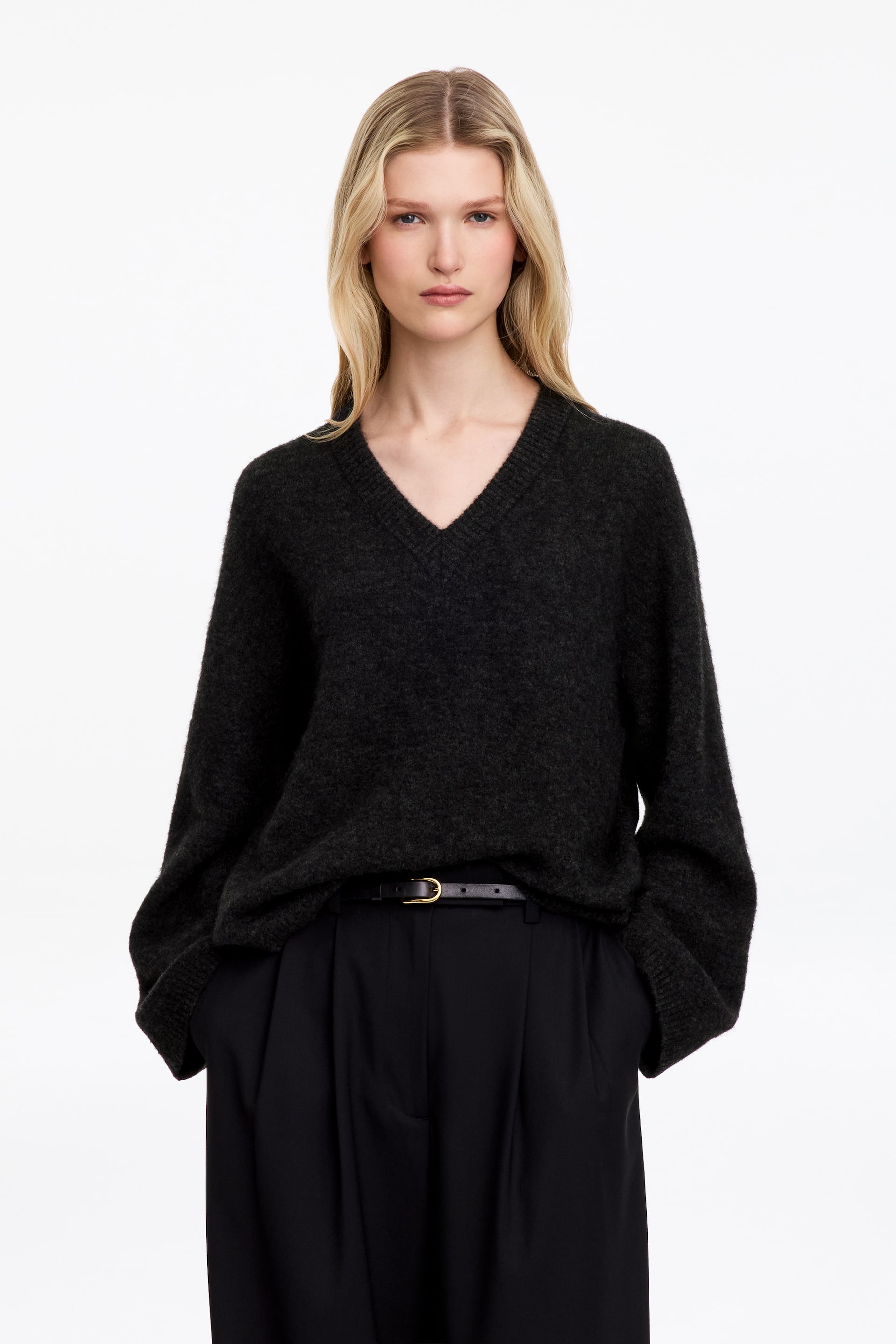Ver imagem maior: Um camisola de malha cinzento-escuro com decote em V, acabamento texturado e corte descontraído está a ser usada pelo modelo. A camisola apresenta mangas compridas e largas e está combinada com umas calças pretas, usada por dentro, com um cinto fino preto.