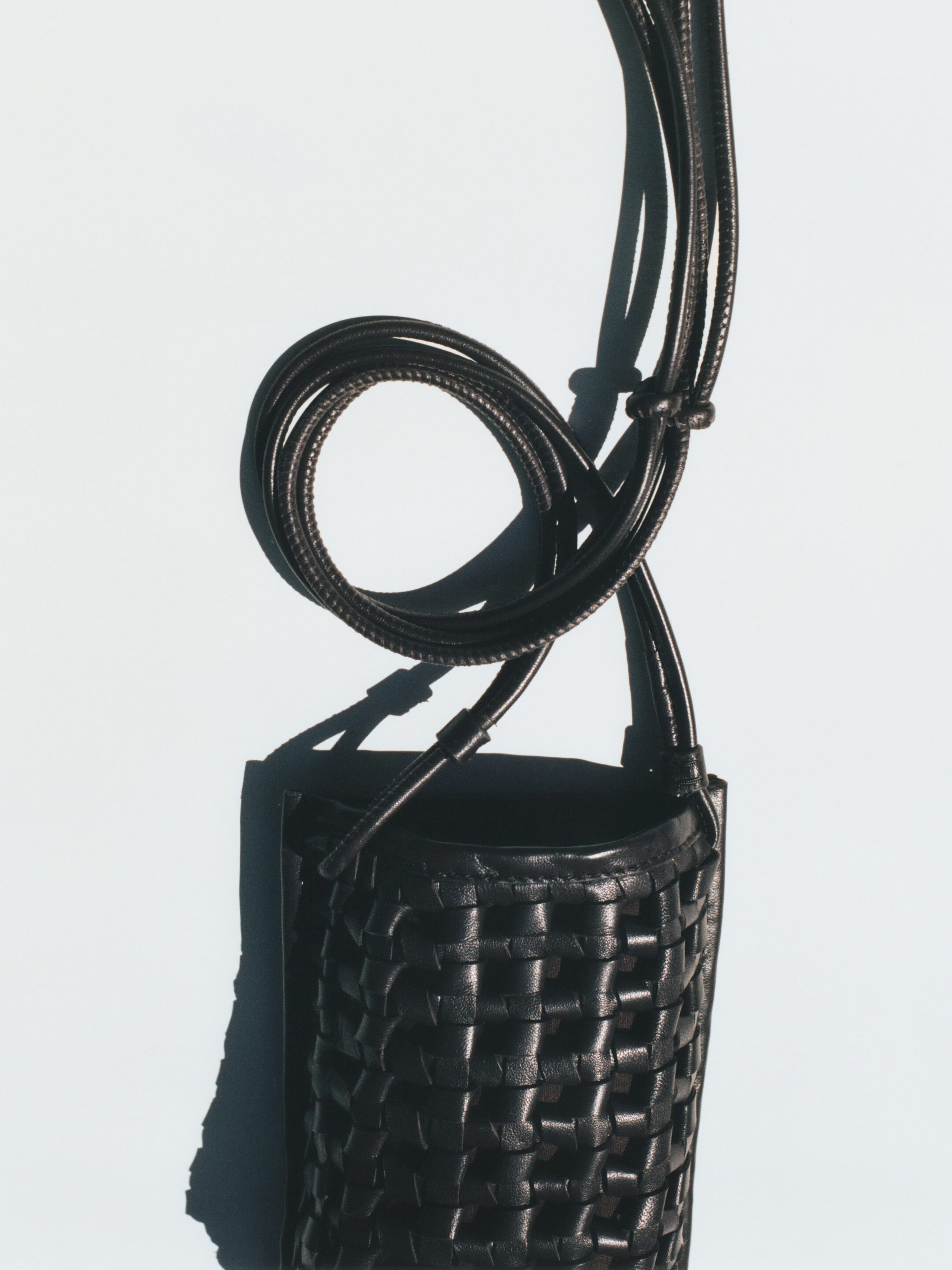 Größeres Bild anzeigen: Eine schwarze Leder-Bucket-Bag hängt vertikal, mit einem geflochtenen Körper und einer offenen Oberseite. Ihr langer, dünner Riemen windet sich über der Tasche und ist am oberen Rand befestigt.