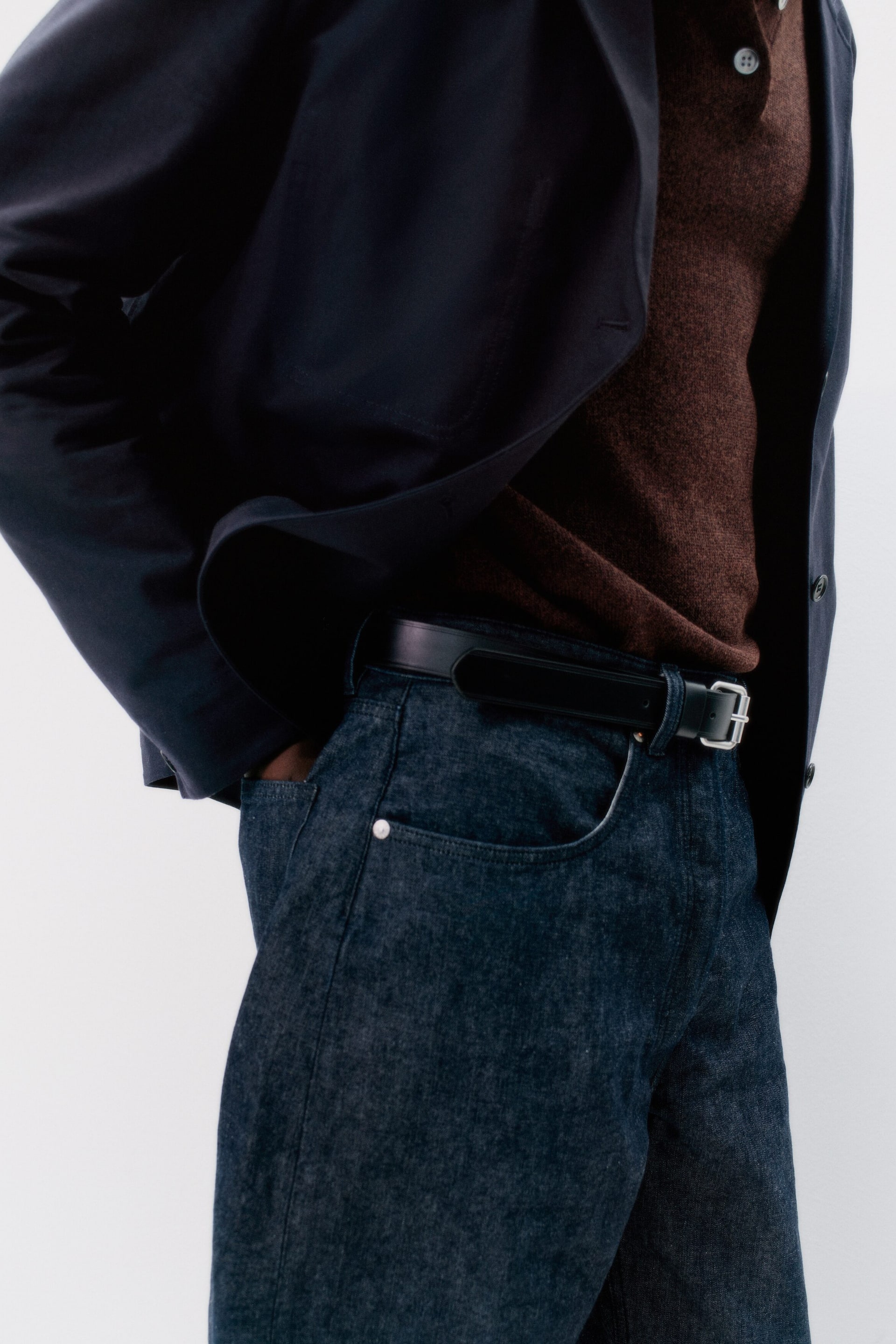 Agrandir l'image: Une veste bleu marine foncé est ouverte, révélant un polo en maille marron avec une patte à deux boutons. Un jean en denim foncé est porté, avec des poches avant et une ceinture en cuir noir avec une boucle argentée, le tout présenté de côté.