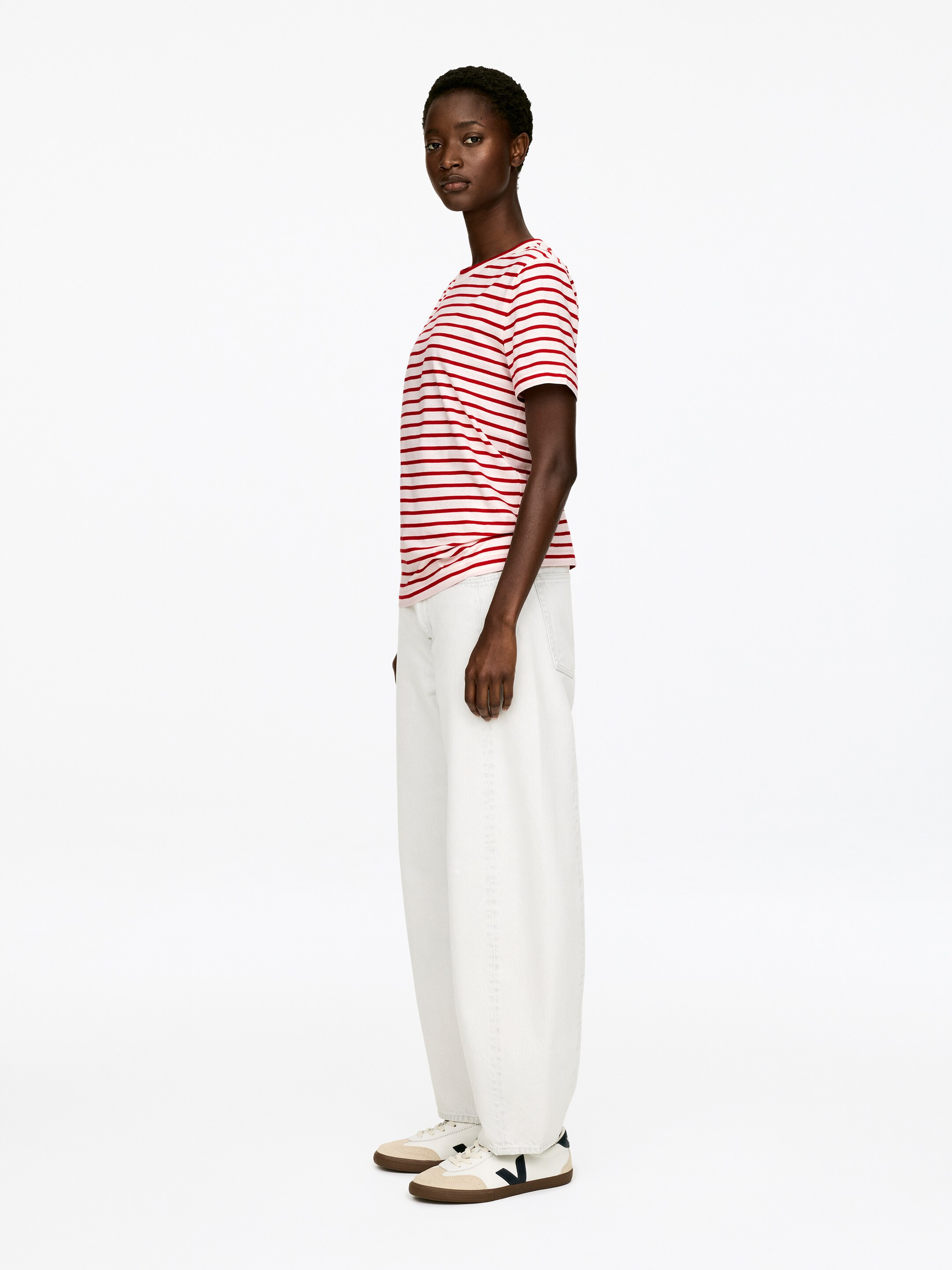 Grotere afbeelding bekijken: Een wit T-shirt met rode horizontale strepen, een ronde halslijn en korte mouwen wordt zijwaarts gepresenteerd. Het is gestyled met off-white, wijde broeken die losjes vallen en lage sneakers met een beige basis, een donkerblauw zijlogo en een bruine zool.
