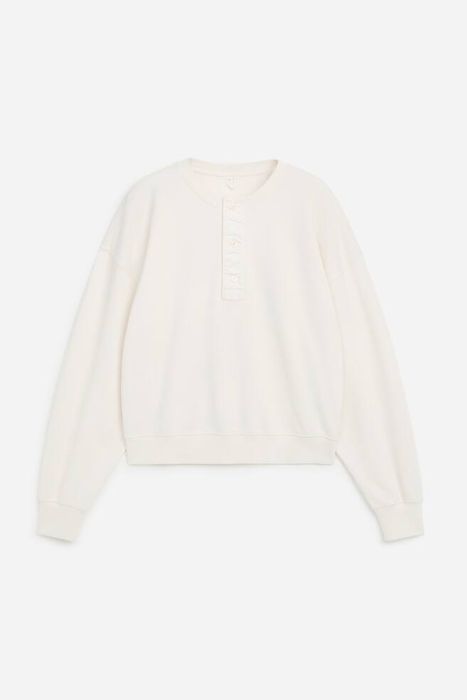 Una felpa henley color crema Ã¨ mostrata frontalmente, con scollo tondo e apertura con quattro bottoni. Presenta una vestibilitÃ  rilassata con spalle scese, maniche lunghe con polsini a coste e orlo a coste. - 1