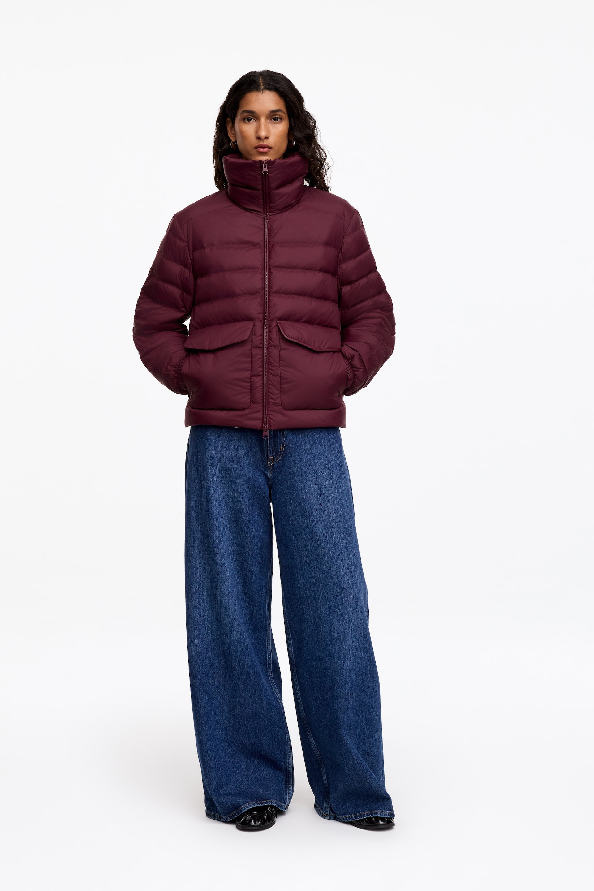 Nagyobb kép megtekintése: A sötét bordó árnyalatú Re:Down Padded Jacket egy vízszintesen steppelt pufi kabát, magas állógallérral, elülső cipzárral és fedeles zsebekkel. A modellen látható, aki a kezét a zsebébe dugva viseli, és bő szárú kék farmerrel van stylingolva.