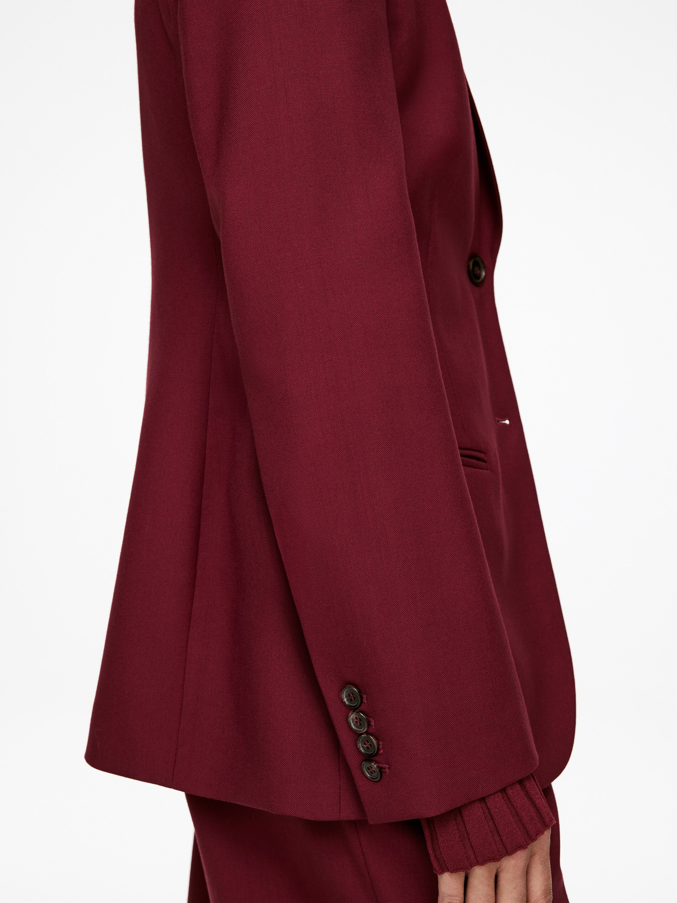Visualizza immagine più grande: Un blazer bordeaux è esposto da un'angolazione laterale, mettendo in mostra la sua vestibilità su misura. La sua manica lunga presenta quattro bottoni scuri sul polsino, mentre un polsino a coste di un indumento coordinato si estende da sotto. Un bottone scuro è anche visibile sulla chiusura frontale del blazer.