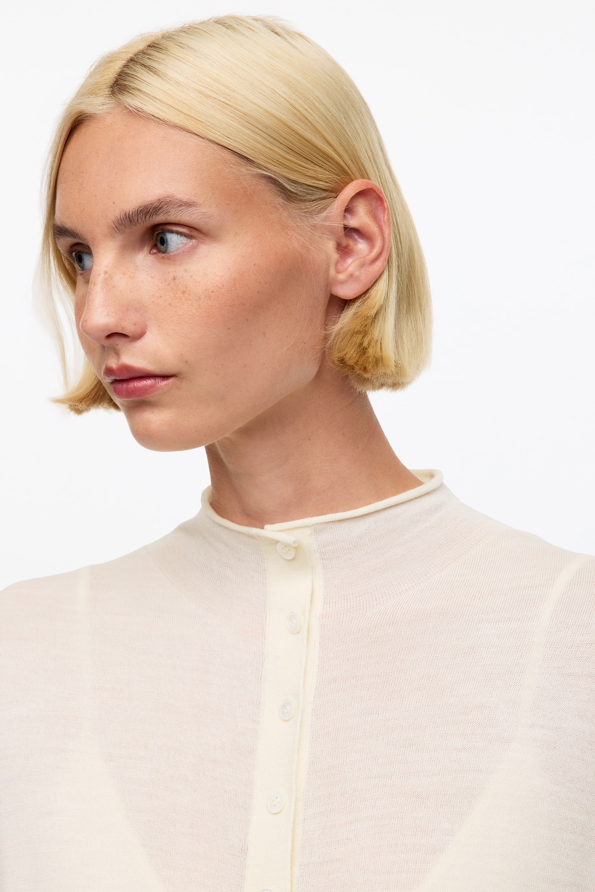 Agrandir l'image: Un cardigan crème en maille fine à boutonnage sur le devant est présenté sous un léger angle avant droit, mettant en valeur son délicat col bande et sa patte de boutonnage. La maille ajourée laisse entrevoir discrètement un sous-vêtement en dessous.