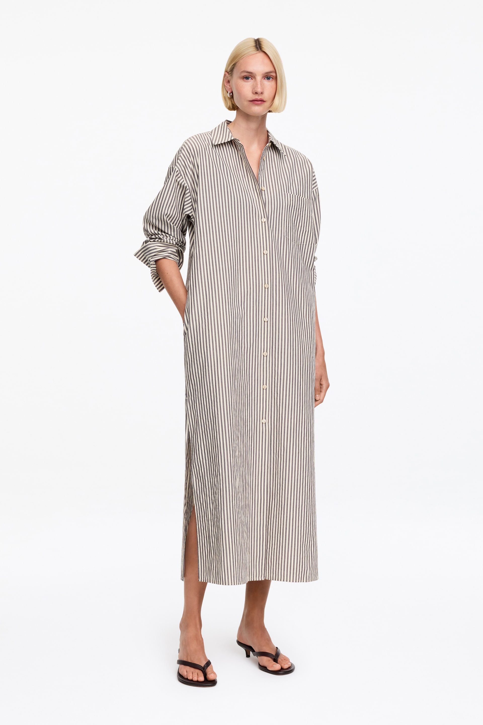 Nagyobb kép megtekintése: Egy függőlegesen csíkos ingruha látható elölről, amely tompa olíva-barna és törtfehér csíkokkal rendelkezik, hegyes gallérral és teljes hosszúságú gombos záródással. Hosszú ujjakkal tervezték, amelyek a mandzsettáknál fel vannak hajtva, bal oldalon egy mellzseb található, és oldalsó hasítékokkal rendelkezik, amelyek a maxi hosszúságú szegélytől indulnak.