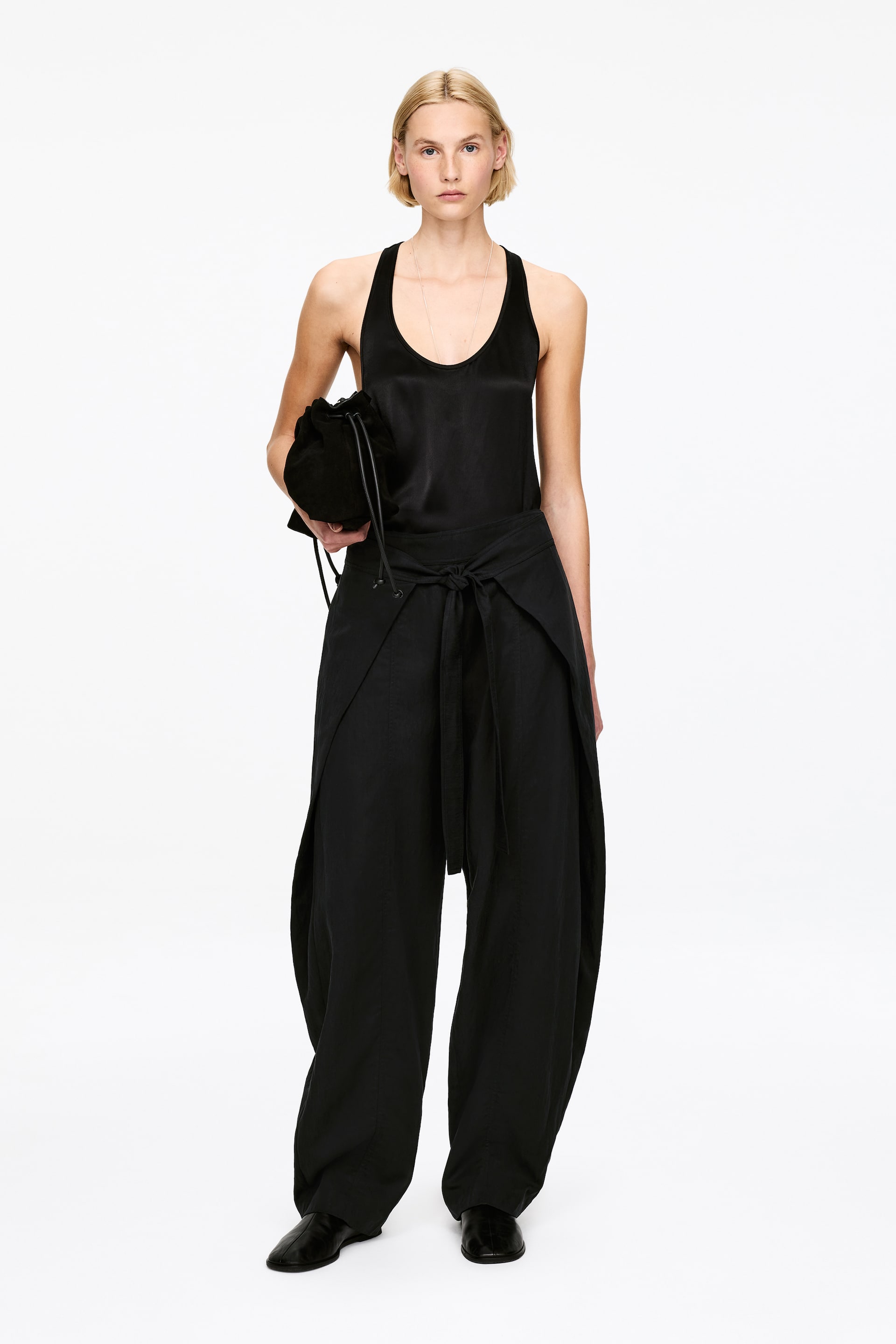 Vezi imaginea mai mare: Un maieu negru din satin cu spatele în stil racerback este asortat cu pantaloni negri voluminoși, în stil wrap, cu o talie cu cordon, prezentat cu fața înainte. Un mic săculeț negru cu șnur este ținut în mână.