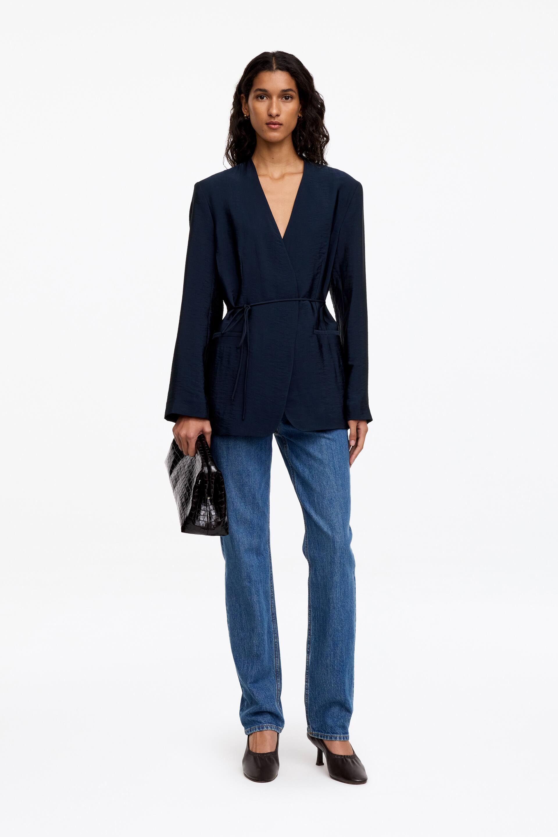 Visualizza l'immagine ingrandita: Un blazer avvolgente senza colletto in blu navy scuro, realizzato in un tessuto liscio con una leggera lucentezza, indossato dalla modella, con un sottile laccio in rouleau annodato in vita e tasche a filo pulite. Il blazer è abbinato a jeans blu a gamba dritta, scarpe nere con tacco e una pochette nera testurizzata tenuta in una mano.