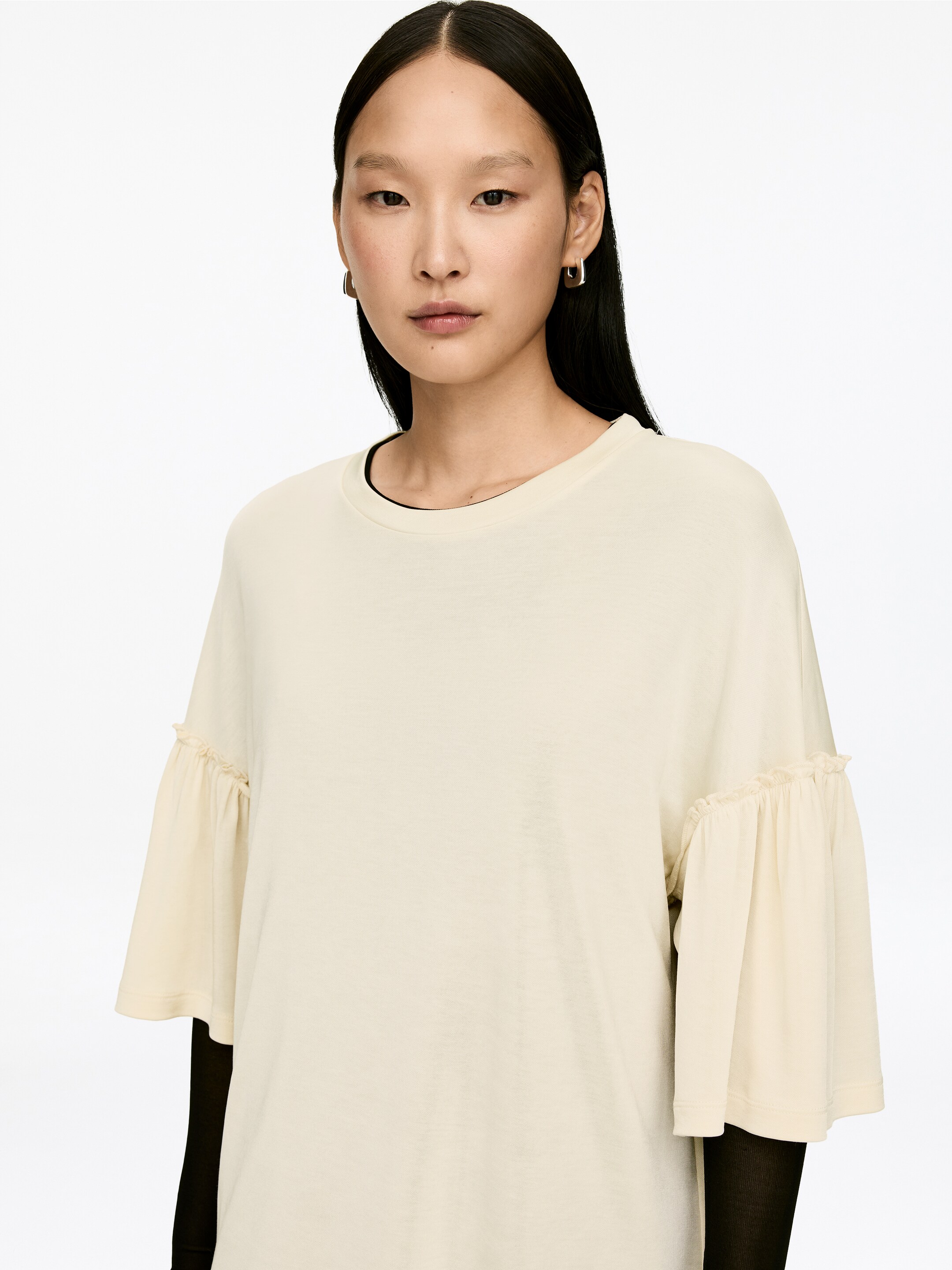 Visa större bild: En krämfärgad T-shirt med ryschade ärmar och normal passform bärs av modellen. En svart långärmad topp är lager under T-shirten.