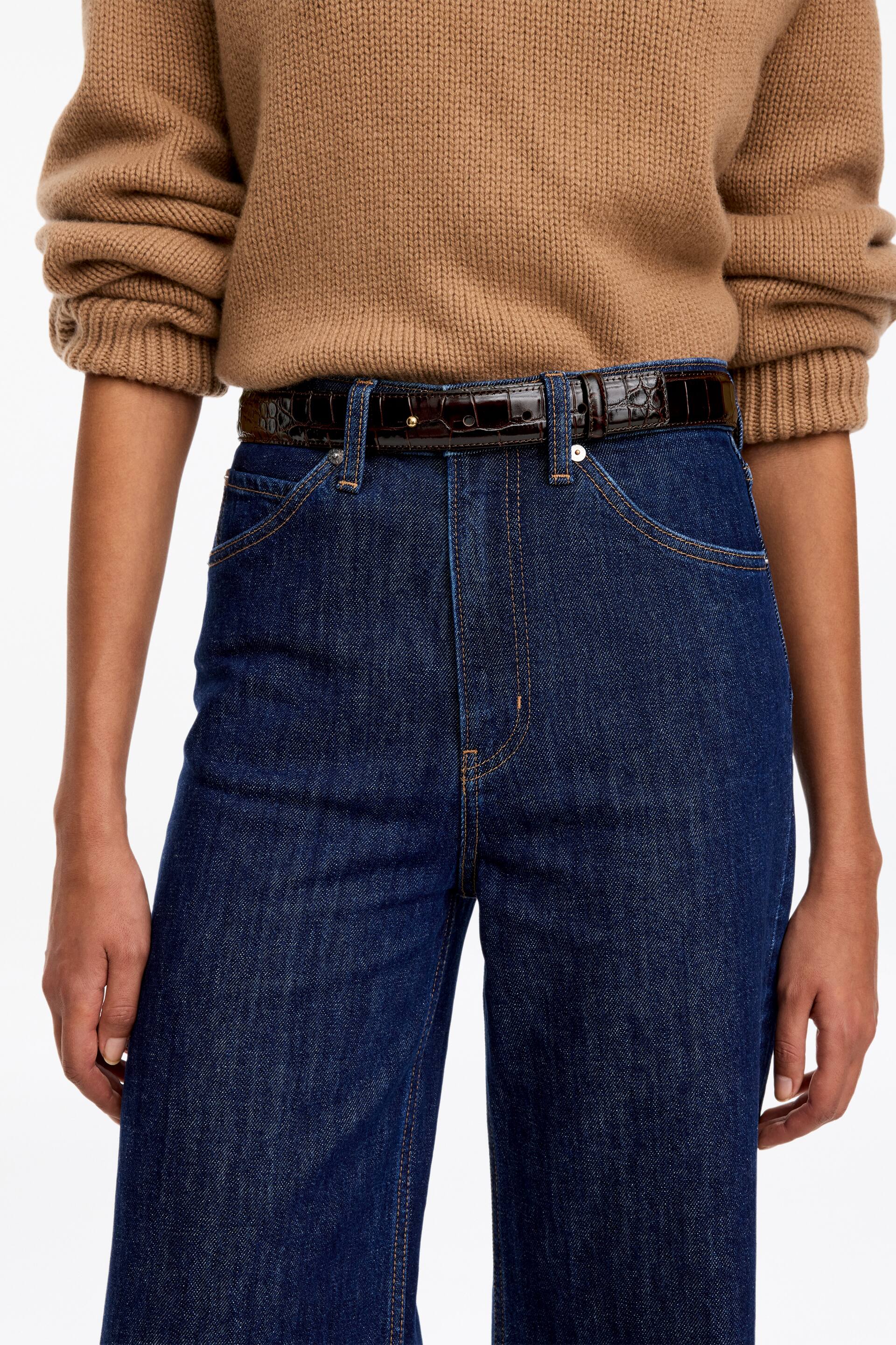 Se større billede: En mørkebrun, blank læderbælte med krokodillemønster bæres gennem bæltestropperne på et par højtaljede blå jeans af modellen. Bæltet har en lille guldfarvet nitte­lukning foran og er stylet med en brun striktrøje, der er stoppet ned.