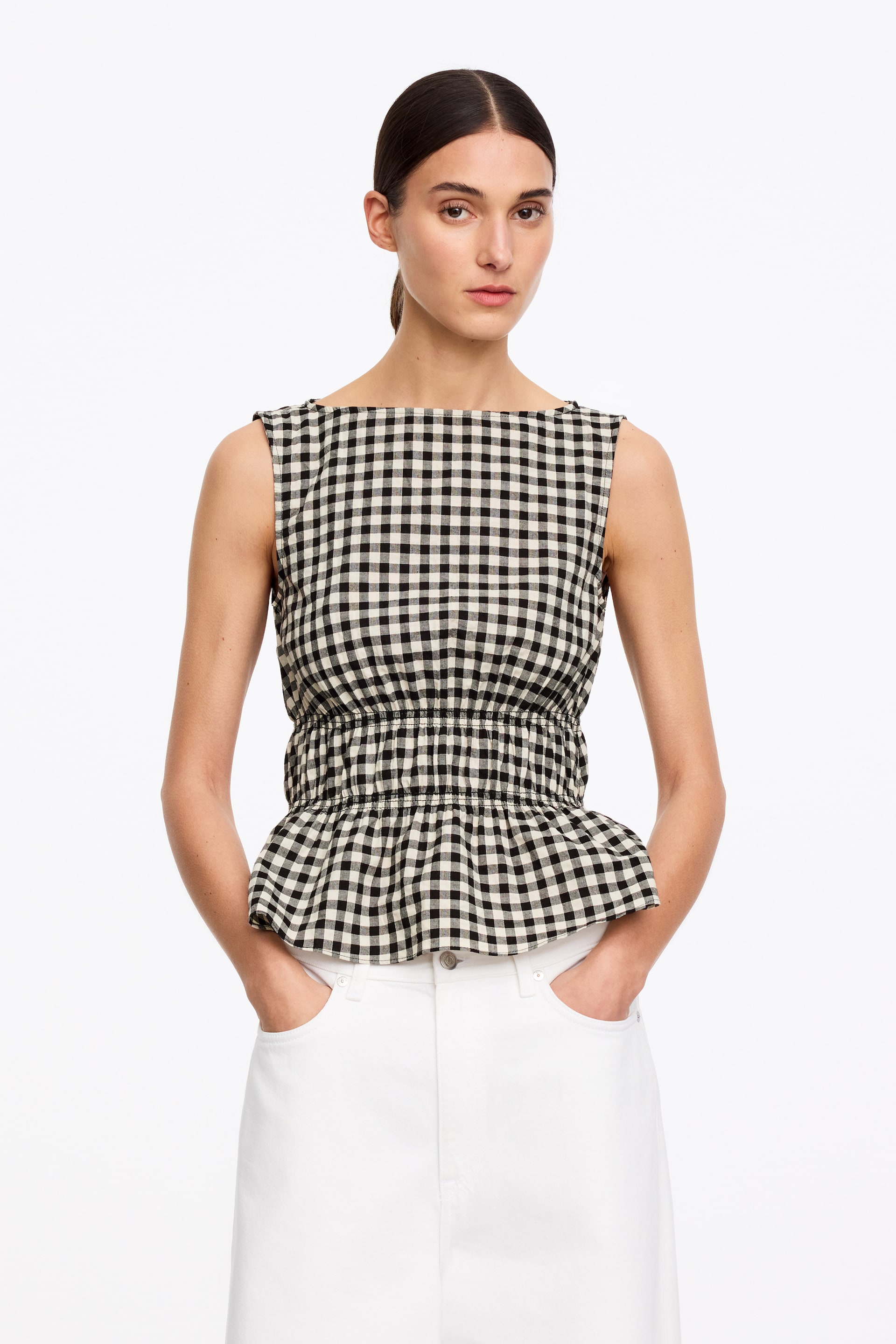 View larger image: A black and white gingham sleeveless top is showcased from the front, featuring a wide boat neck and a shirred elasticated waist that creates a peplum hem.