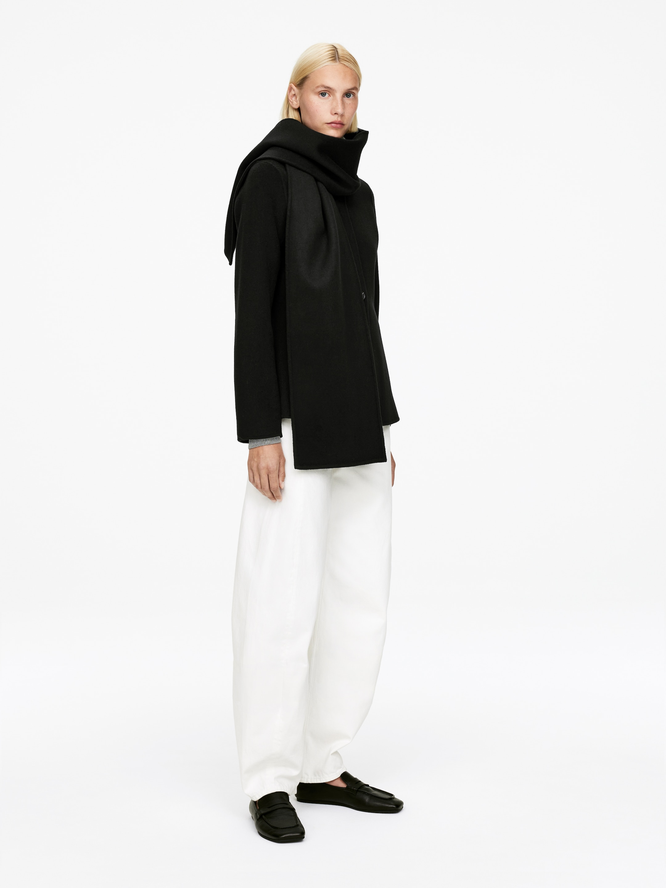 Agrandir l'image: Un manteau noir à coupe décontractée, avec une écharpe intégrée ou attachée, est présenté sous un léger angle latéral. L'écharpe s'enroule autour du cou haut et descend sur le devant, avec une extrémité qui s'étend sur l'épaule et le dos. Le manteau est assorti à un pantalon blanc à jambes larges et des mocassins noirs, et un haut gris clair à manches longues est visible sous les poignets du manteau.