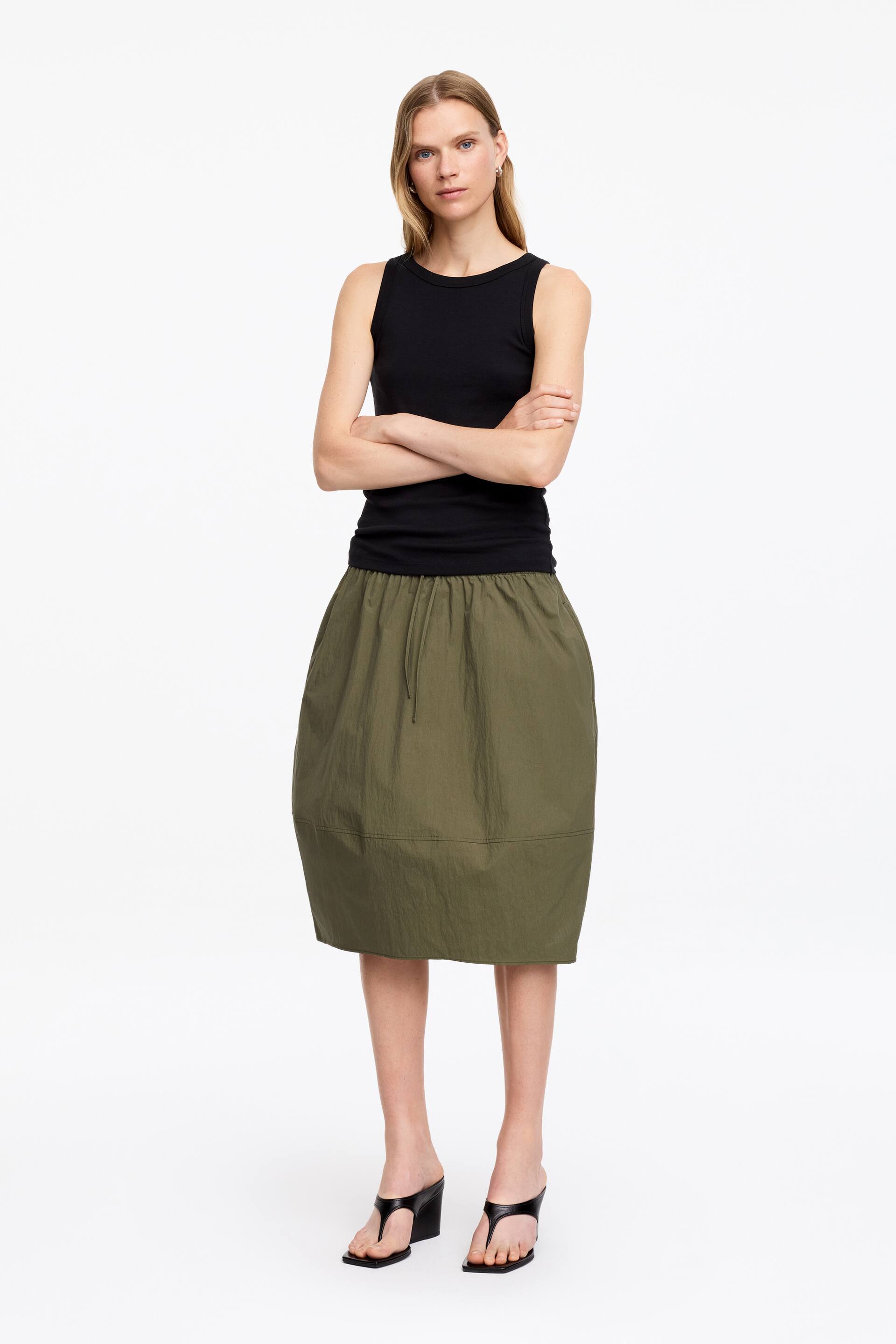 Grösseres Bild ansehen: Ein schwarzes, geripptes Strick-Tanktop mit Rundhalsausschnitt wird frontal präsentiert und mit einem olivgrünen knielangen Rock kombiniert. Der Rock verfügt über einen elastischen Kordelzug in der Taille, eine abgerundete Ballonsilhouette mit horizontaler Naht und wird mit schwarzen Square-Toe-Wedge-Flip-Flop-Sandalen getragen.