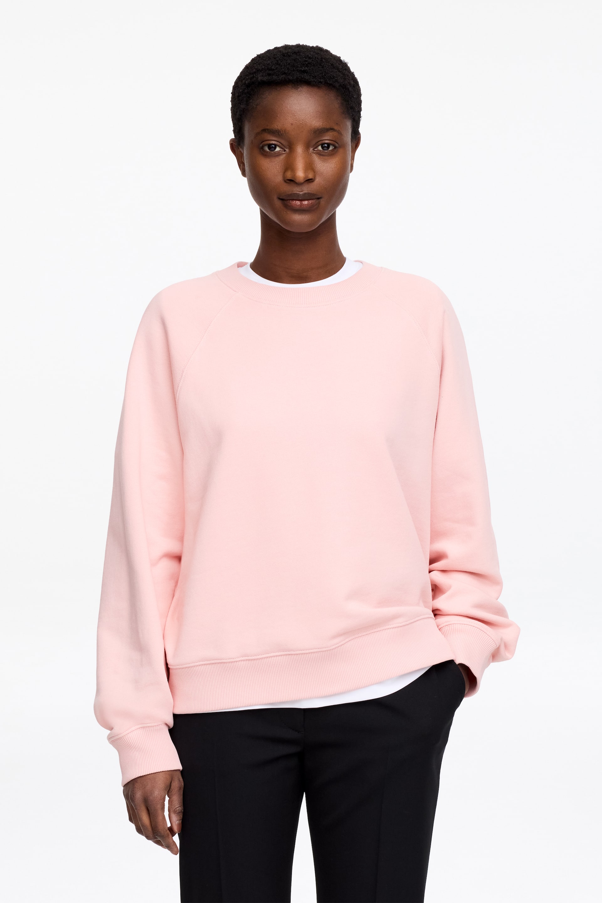 Agrandir l'image: Un sweatshirt rose à col rond est présenté face à l'avant, avec des manches raglan, un col rond côtelé, des poignets côtelés et un ourlet côtelé. Un t-shirt blanc est superposé en dessous, visible au niveau de l'encolure et de l'ourlet, et une main repose dans une poche, tirant doucement le sweatshirt à la hanche.