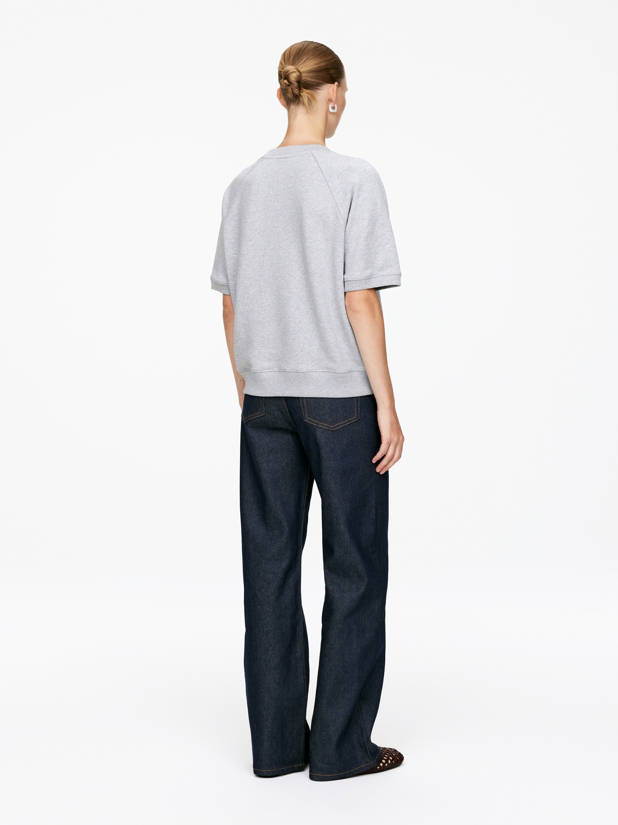 Agrandir l'image: Un sweat-shirt gris à manches courtes est présenté de dos, avec des manches raglan et un ourlet à bande. Il est associé à un jean à jambes larges et lavage foncé, avec des coutures contrastantes visibles.