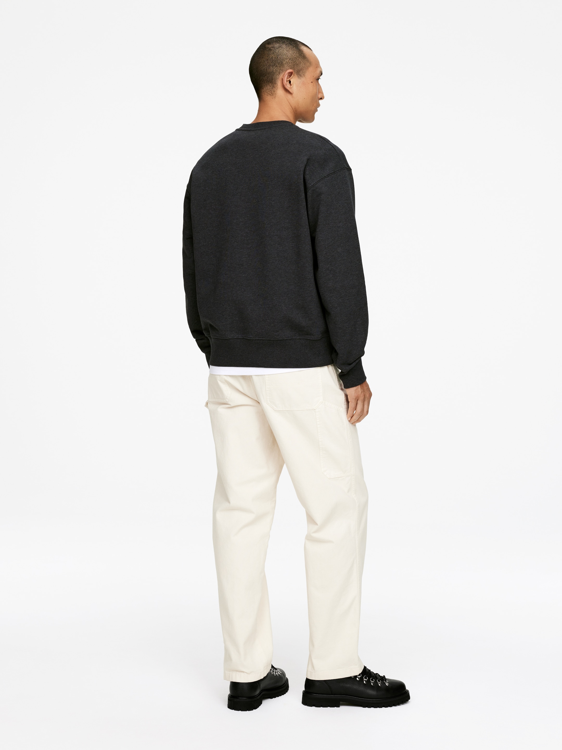 Größeres Bild ansehen: Ein dunkelgraues Sweatshirt mit Rundhalsausschnitt wird getragen, von hinten und von der Seite betrachtet, mit einer entspannten Passform, gerippten Bündchen und einem gerippten Saum, über einem weißen T-Shirt. Hellcremefarbene Utility-Hosen zeigen eine gerade Passform und aufgesetzte Taschen, kombiniert mit schwarzen Schnürschuhen, die dicke Profilsohlen aufweisen.