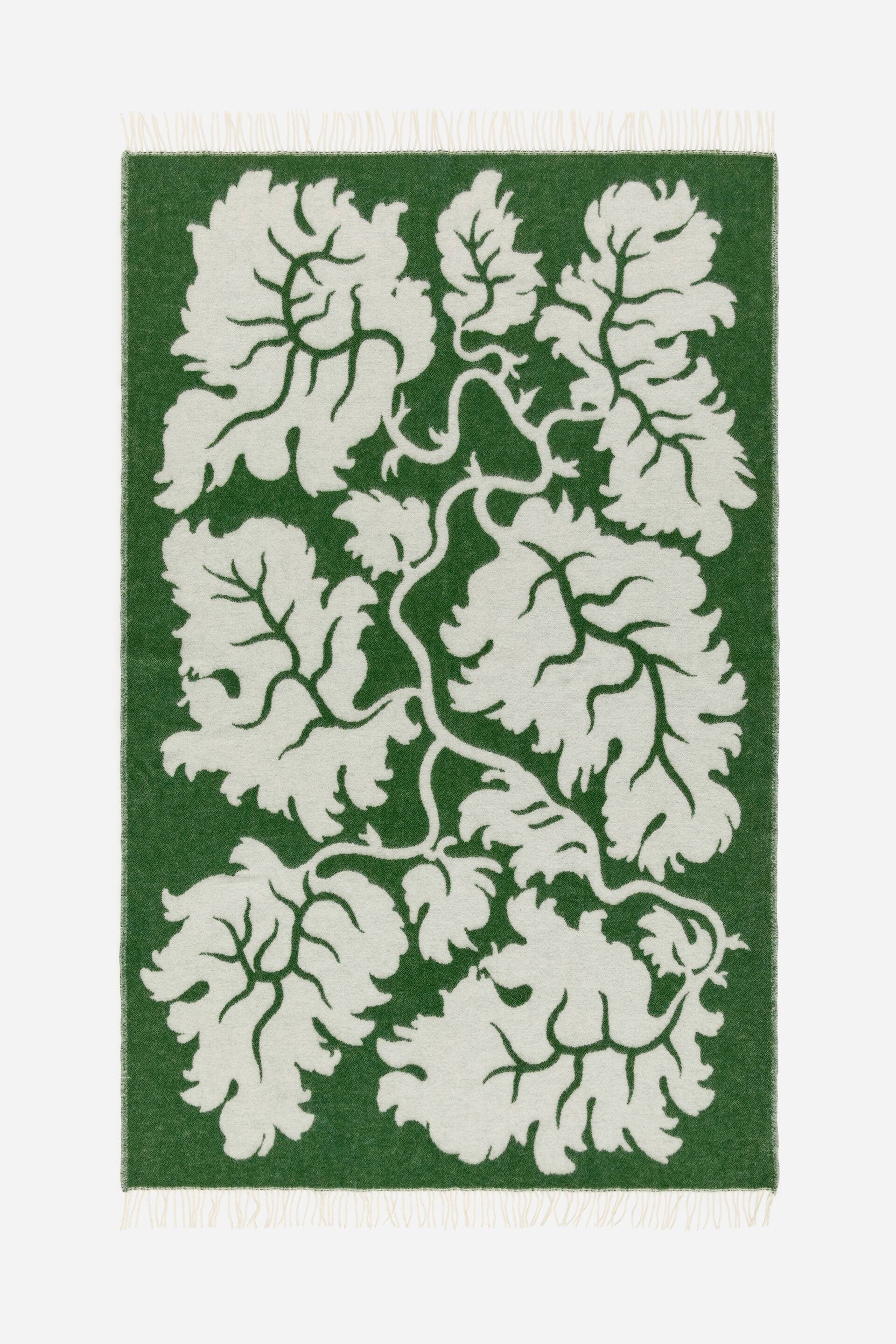 Agrandir l'image: Un plaid rectangulaire arbore un grand motif botanique blanc cassé de feuilles stylisées et de lianes sinueuses sur un fond vert foncé, avec des franges crème finissant les bords supérieur et inférieur.