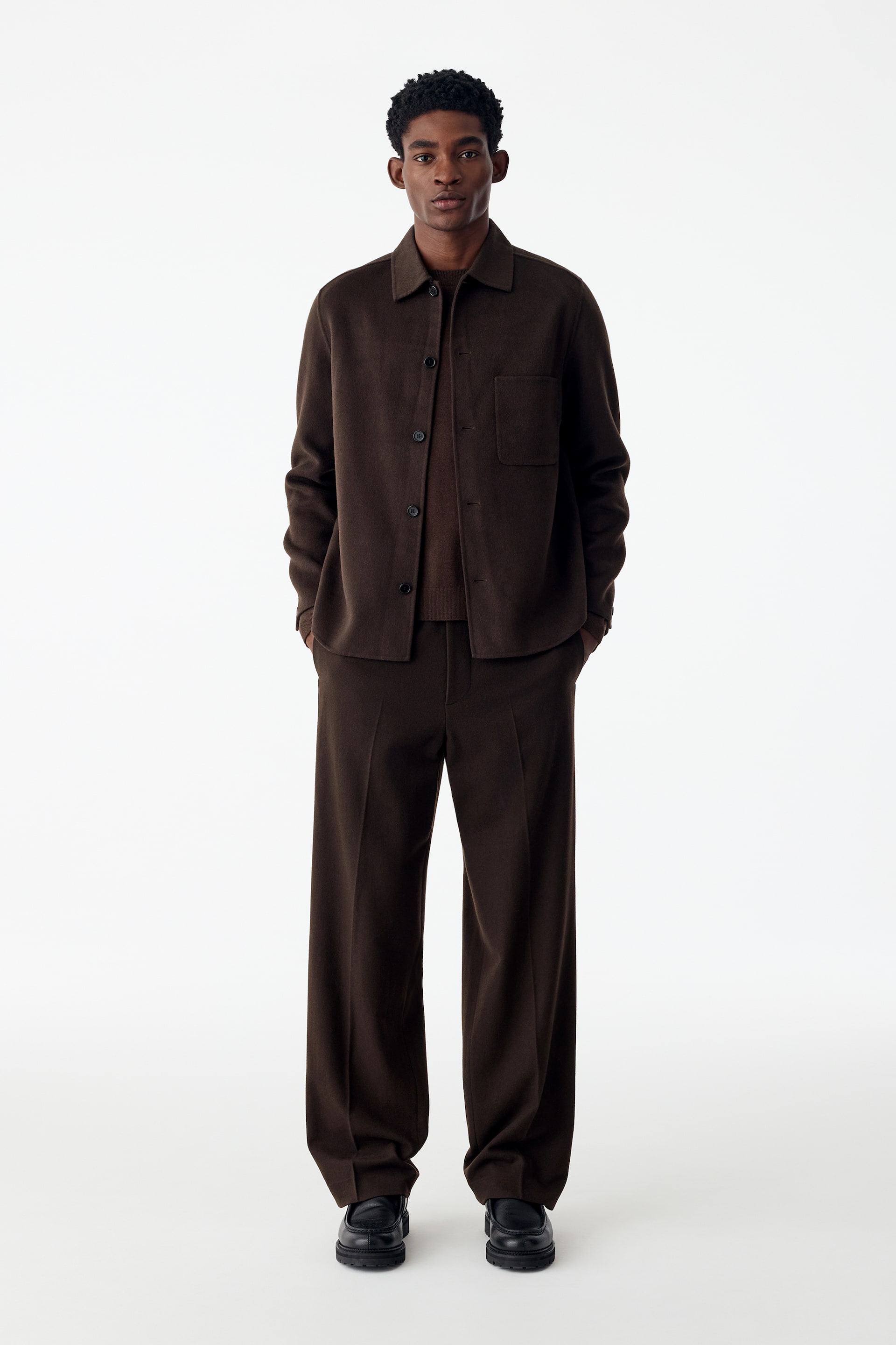 Ver imagem maior: A modelo veste uma overshirt em mistura de lã castanho-escura, com gola clássica, fecho frontal por botões e um único bolso de chapa no peito. A overshirt estruturada é usada sobre um top a condizer e combinada com calças de perna larga a condizer.
