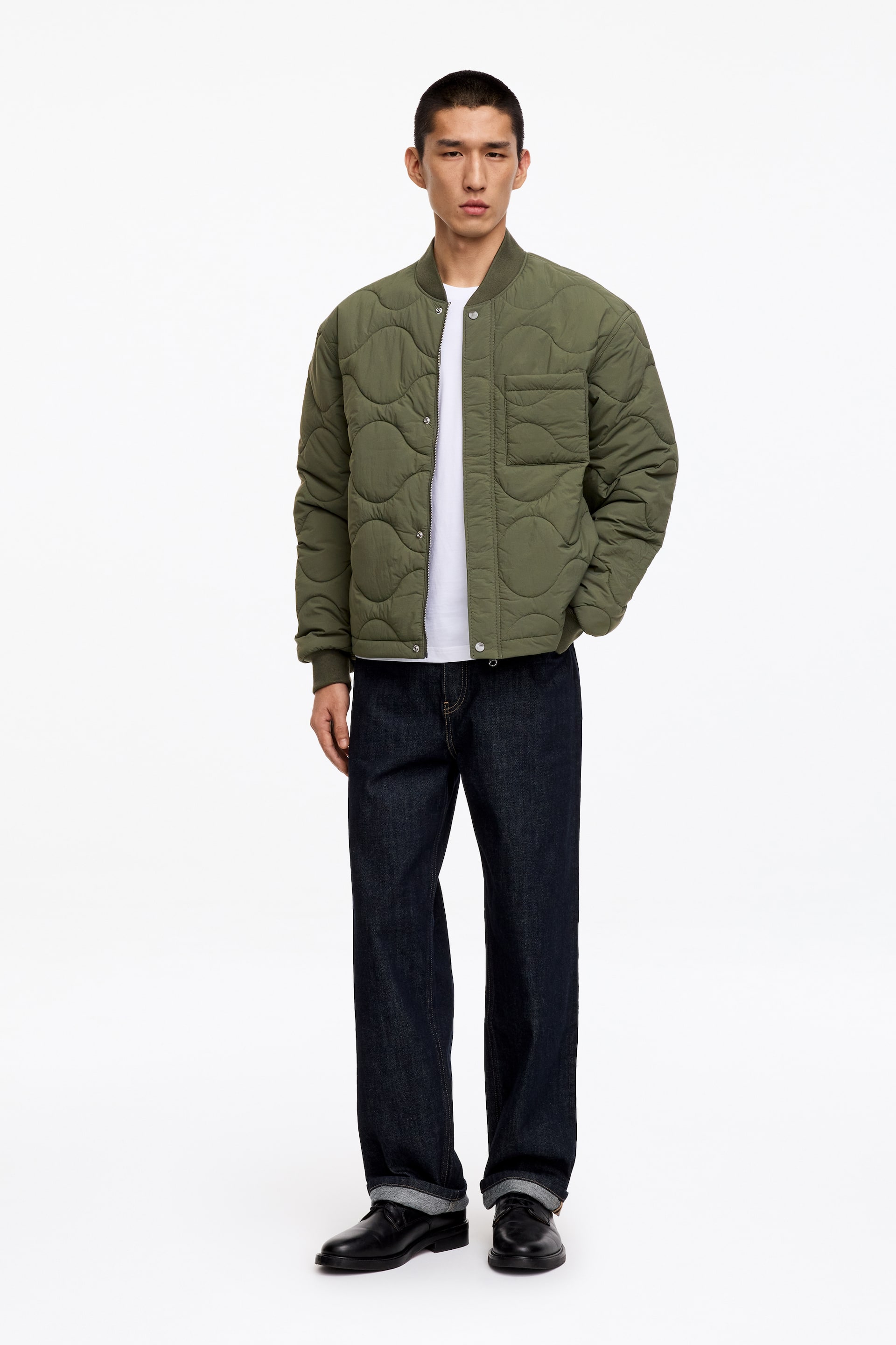 Größeres Bild anzeigen: Eine olivgrüne, leichte, wattierte Innenjacke mit einem gesteppten Zwiebelmuster wird vom Model über einem weißen T-Shirt getragen. Die Jacke hat eine offene Frontleiste mit sichtbaren Druckknöpfen, eine einzelne Brusttasche und gerippte Abschlüsse an den Bündchen und am Saum, wobei eine Hand des Models in einer Jackentasche ruht.