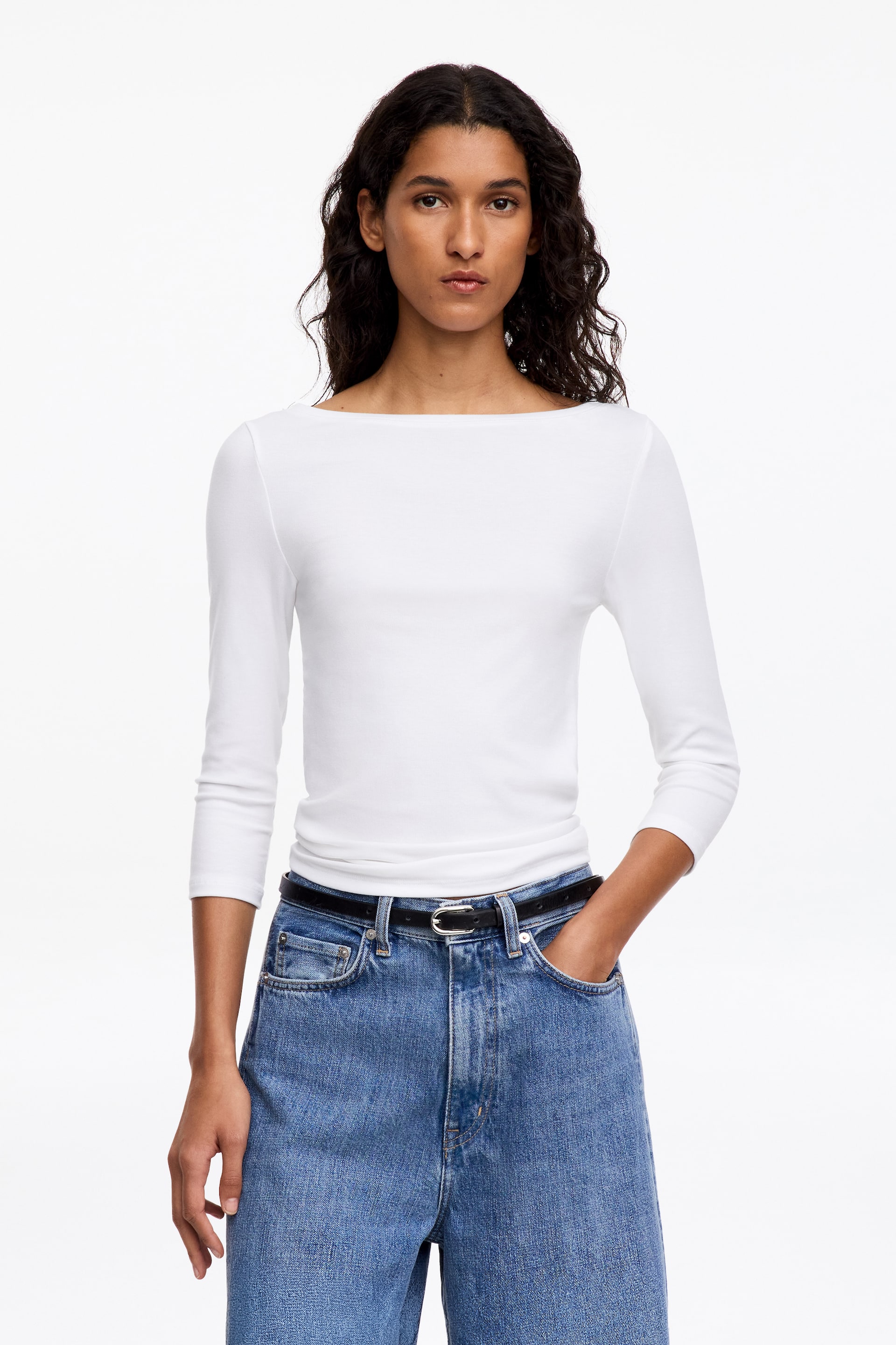 Visa större bild: En vit topp med slim fit som har båtringning och trekvartsärmar bärs av modellen. Toppen är instoppad i blåa jeans i denim, som hålls på plats med ett svart bälte.