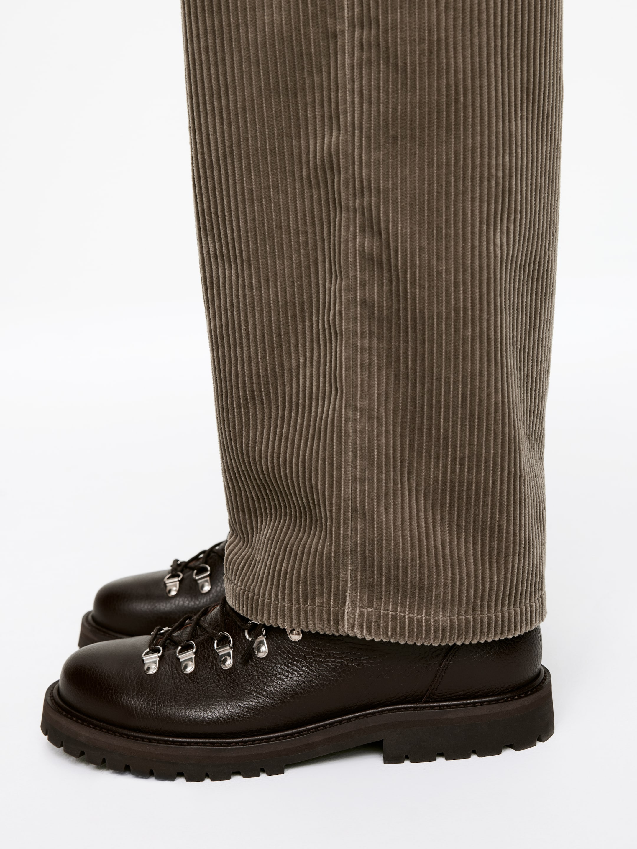 Agrandir l'image: Un pantalon en velours côtelé brun atténué tombe droit sur une paire de bottes en cuir grainé brun foncé. Les bottes présentent des semelles épaisses et un laçage avec des anneaux en D de couleur argentée, vues sous un léger angle de face.