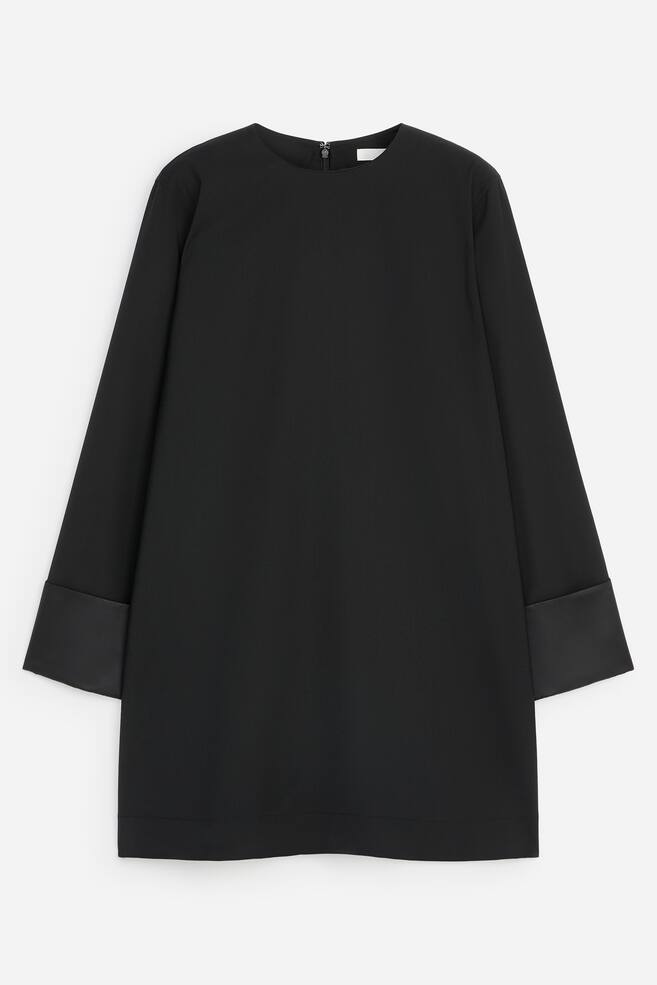 A black shift dress is shown from the front, featuring a crew neckline and long sleeves with wide, contrasting satin cuffs. The dress presents a loose, straight-cut silhouette with a matte finish on the main body and a small zip detail visible at the back of the collar. - 1
