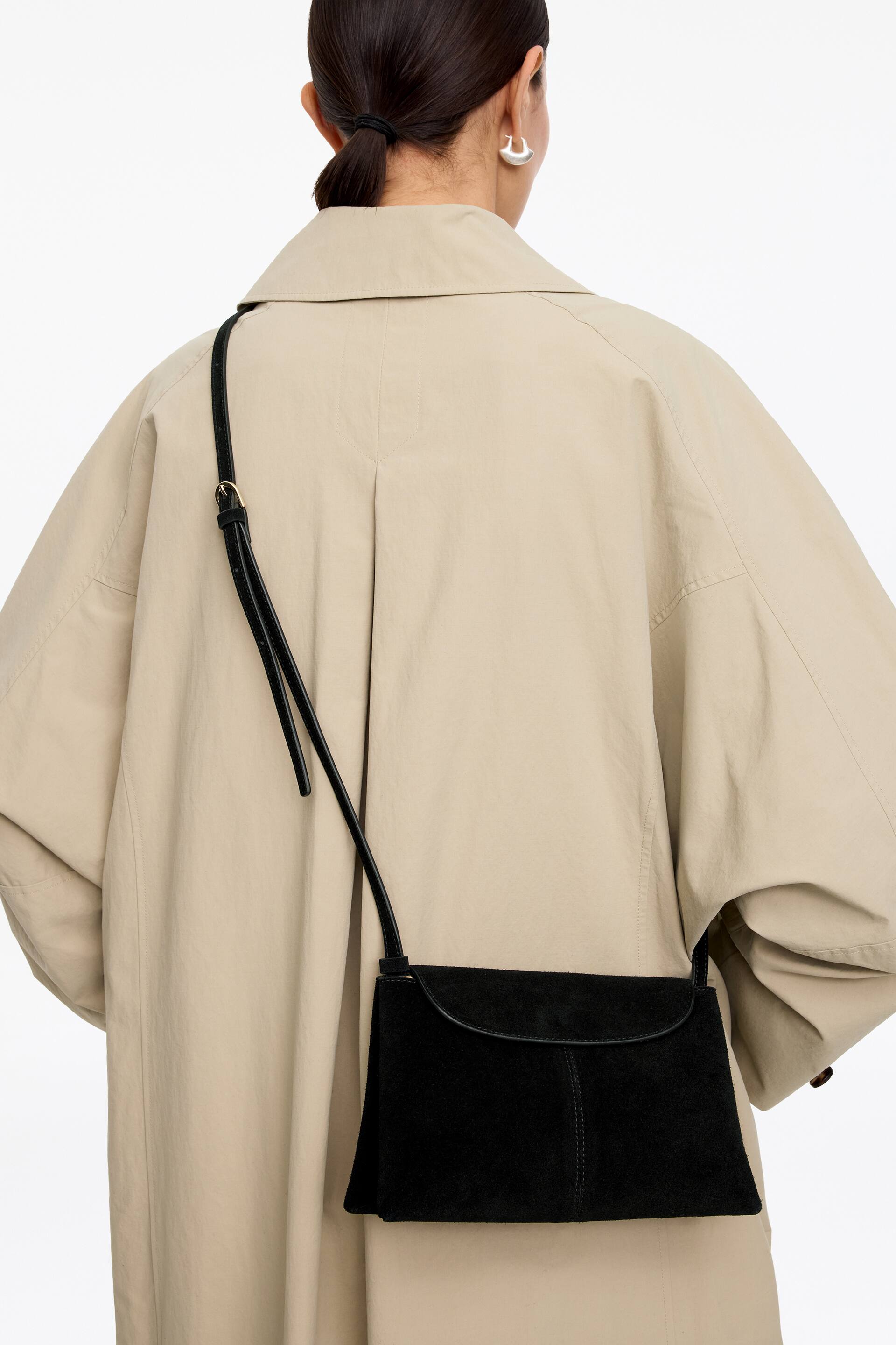 Näytä suurempi kuva: Musta mokkanahkainen crossbody-laukku on käytössä, sen etuläppä kaarevalla reunalla eteenpäin. Ohut säädettävä hihna on näkyvissä, samoin pystysuora sauma etupaneelissa.