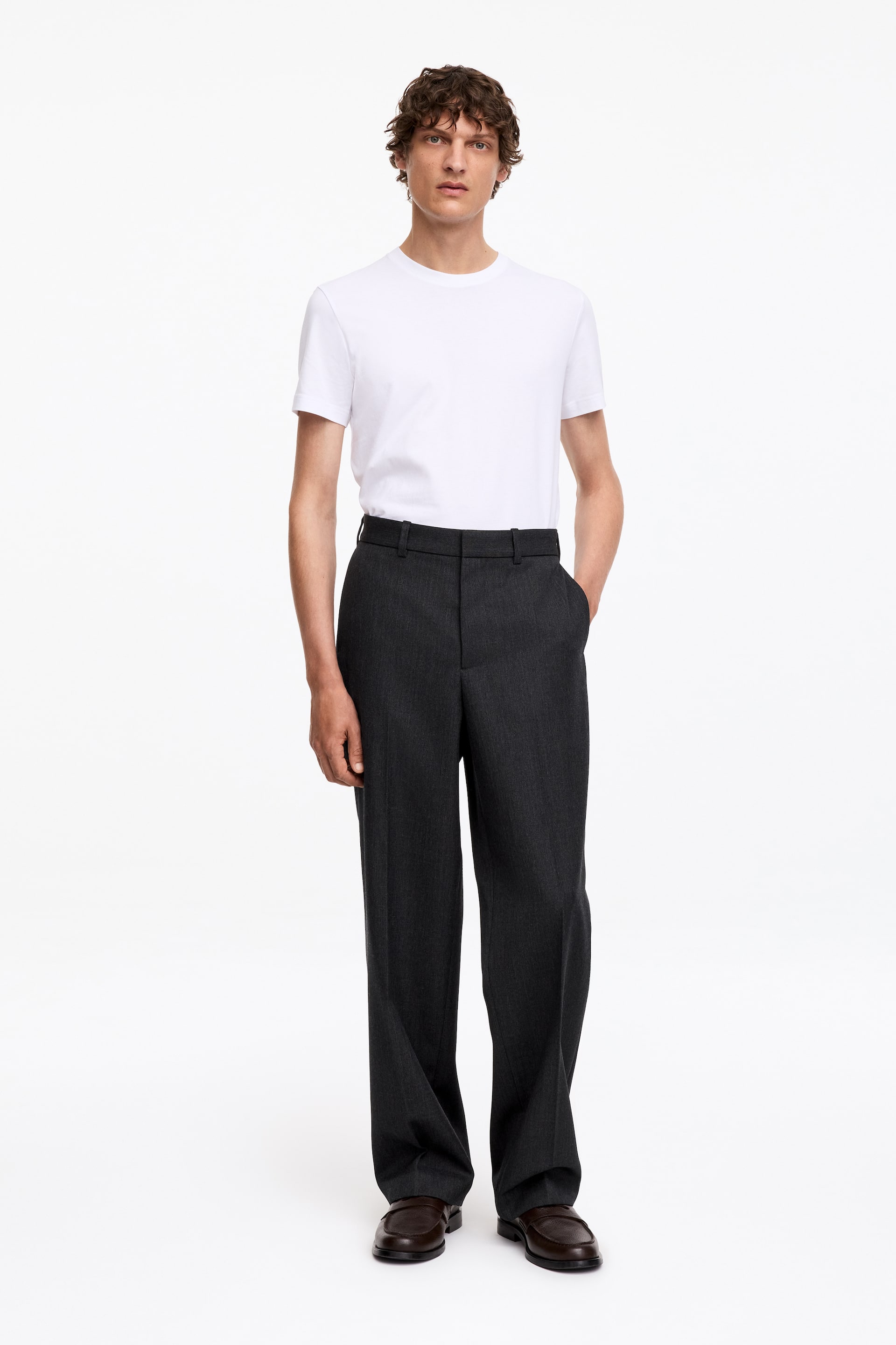 Vezi imaginea mai mare: Pantaloni relaxați, gri închis, cu dungi călcate și găici pentru curea, purtați de model. Pantalonii sunt asortați cu un tricou alb băgat în pantaloni și pantofi din piele maro închis, în timp ce modelul stă cu o mână sprijinită într-un buzunar lateral.