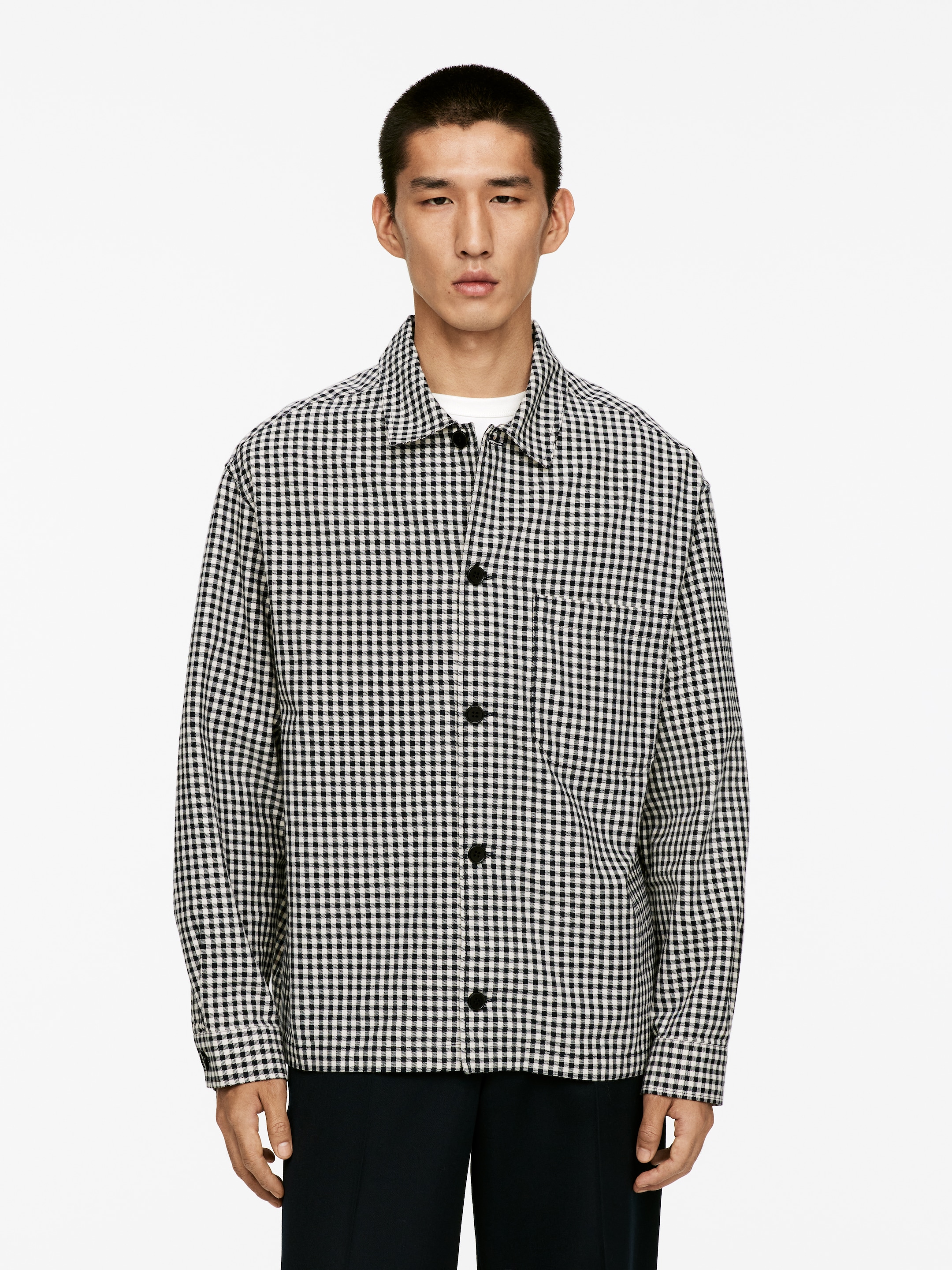 Grösseres Bild ansehen: Ein schwarz-weißes Gingham-Hemd wird präsentiert, das einen spitzen Kragen, schwarze Knöpfe an der Vorderseite und eine einzelne aufgesetzte Tasche auf der linken Brust aufweist. Ein weißes T-Shirt mit Rundhalsausschnitt ist unter dem offenen Kragen sichtbar.