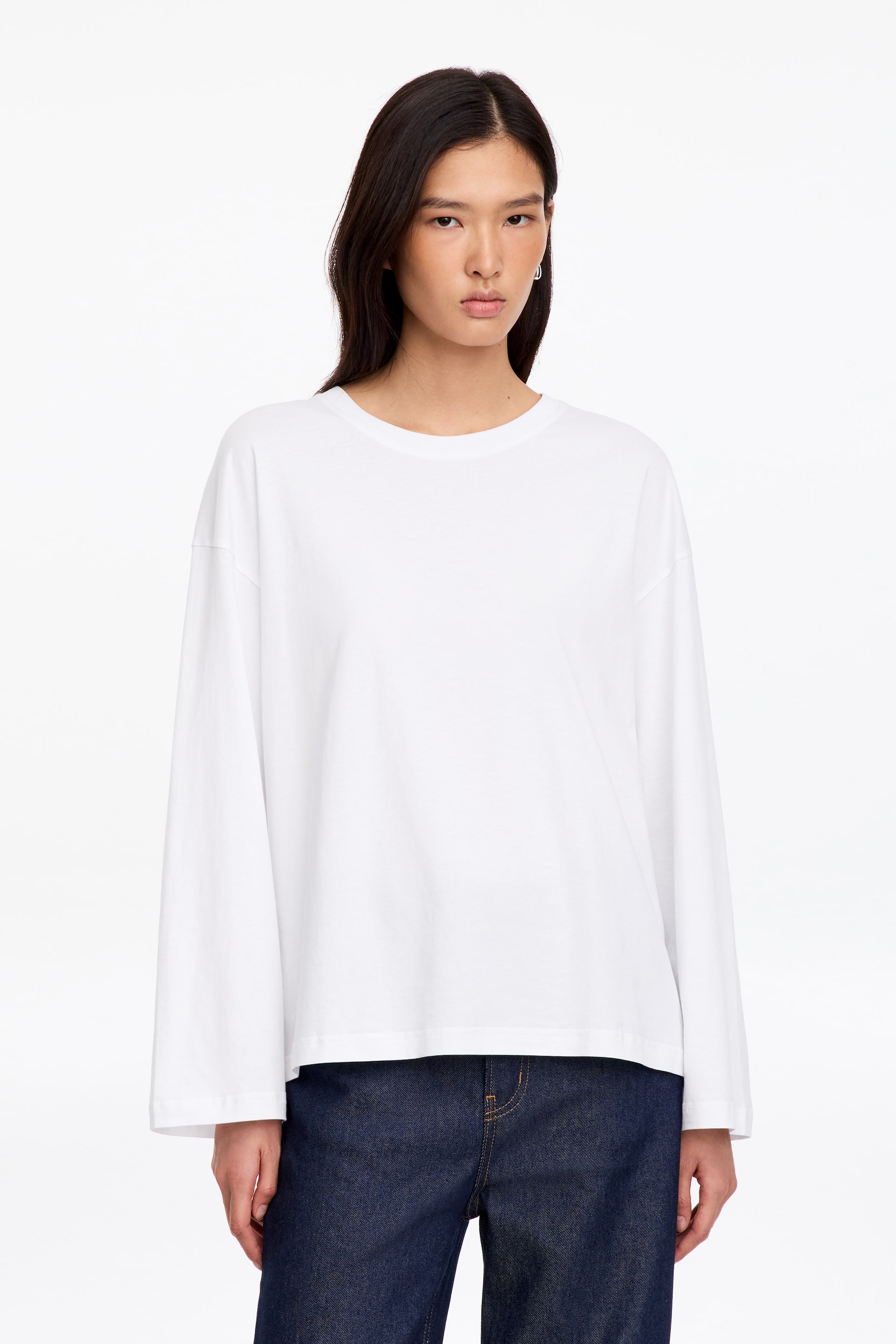 Visa större bild: Modellen bär en vit långärmad T-shirt med avslappnad passform, rund halsringning och sänkta axlar. T-shirten är stylad med mörkblå denimjeans.