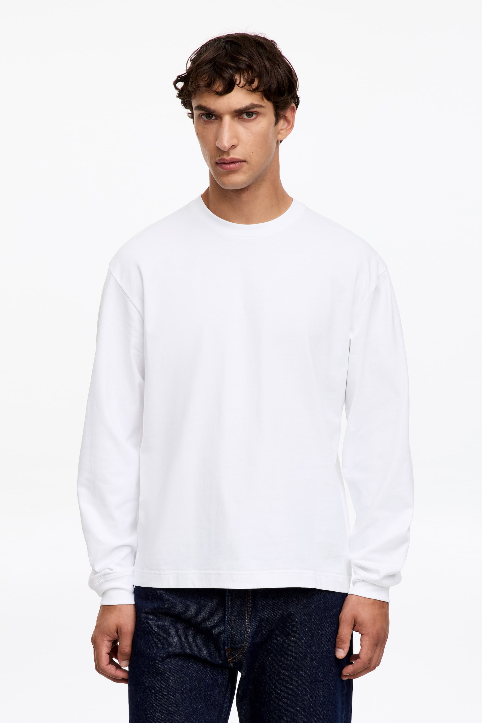 Visualizza immagine più grande: Una T-shirt in cotone bianco a maniche lunghe presenta una vestibilità morbida con spalle scese, polsini a costine e girocollo a costine. La modella indossa la T-shirt con jeans blu scuro.