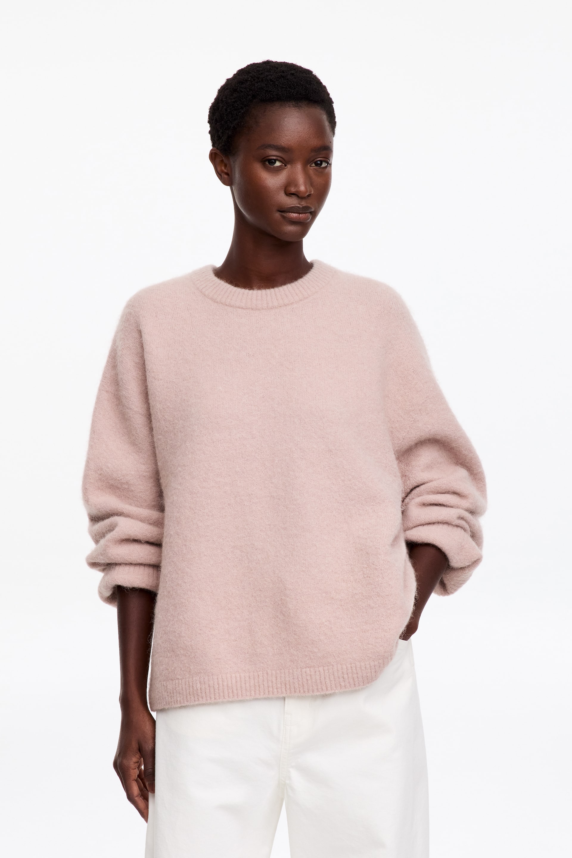 Se større billede: En lyserød crewneck-sweater med blød, fluffy tekstur bæres af modellen. Sweateren har ribkant ved halsudskæringen og forneden samt løstsiddende ærmer skubbet op på underarmene og er stylet med hvide bukser.