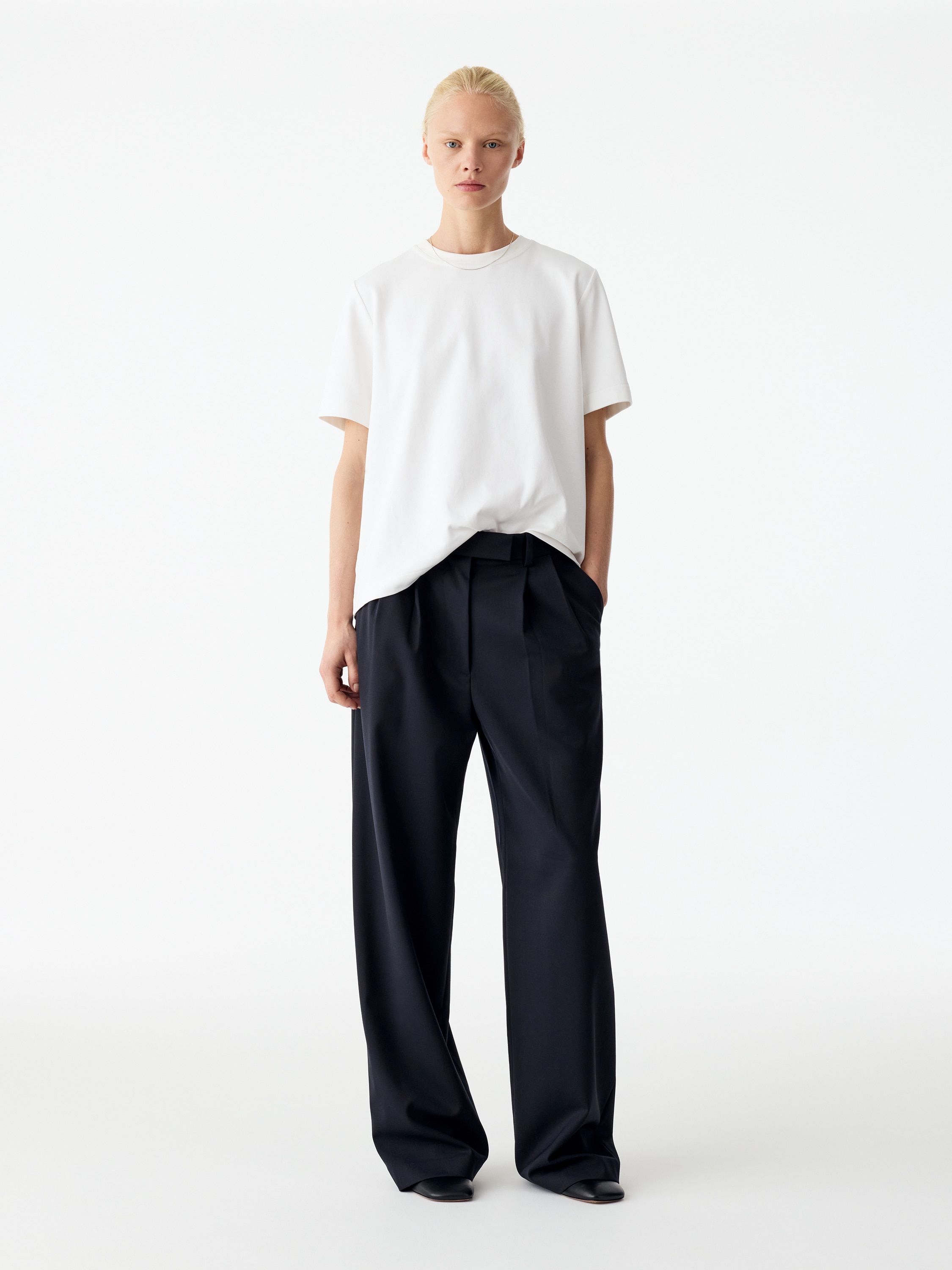 Questi pantaloni blu navy scuro, mostrati da un'angolazione frontale, presentano una silhouette ampia e rilassata con pieghe frontali strutturate. Sono dotati di vita con passanti per cintura, tasche laterali a filo e una chiusura frontale pulita e nascosta.