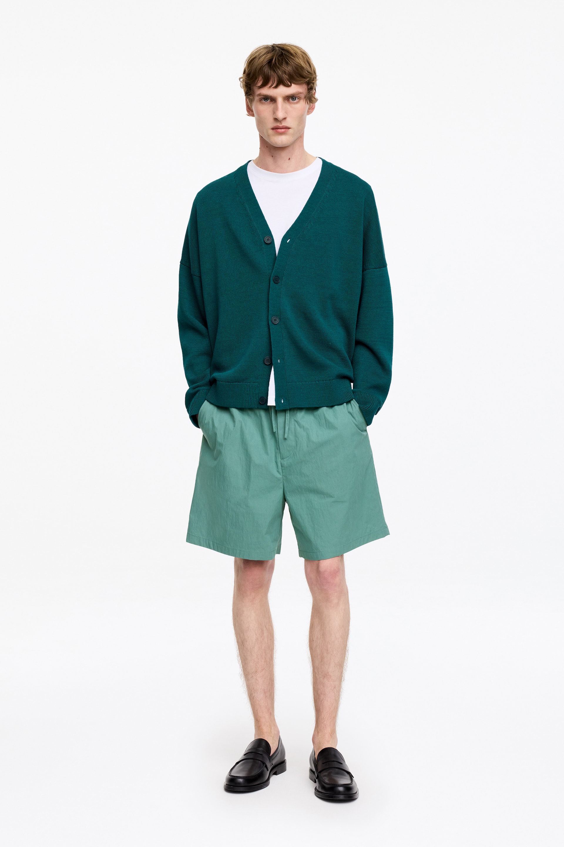Grotere afbeelding bekijken: Een diep groene V-hals cardigan wordt gedragen door het model, gelaagd over een wit t-shirt. Deze cardigan met een ontspannen pasvorm heeft donkere knopen aan de voorkant en lange mouwen.
