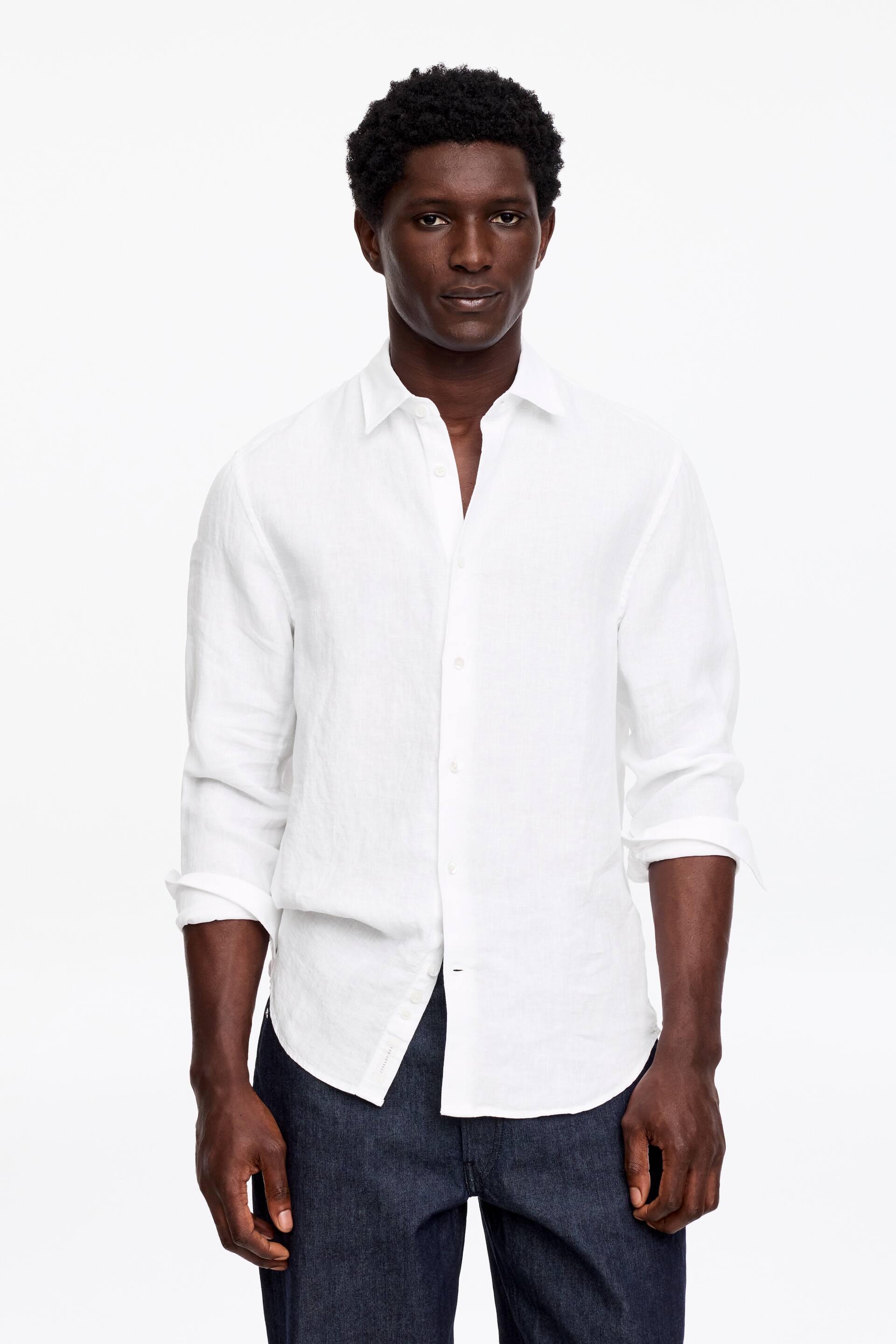 Visa större bild: En vit linneskjorta presenteras framifrån, med en spridd krage där de översta knapparna är öppna, långa ärmar uppkavlade till underarmarna och en böjd fåll delvis instoppad i mörka jeans.