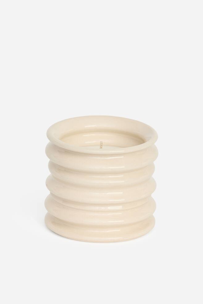 Una vela aromÃ¡tica descansa en el interior de un recipiente de gres de color crema y acabado brillante, con estrÃ­as horizontales apiladas. La vela de una sola mecha se muestra dentro del recipiente cilÃ­ndrico, que presenta un borde superior ligeramente acampanado. - 1