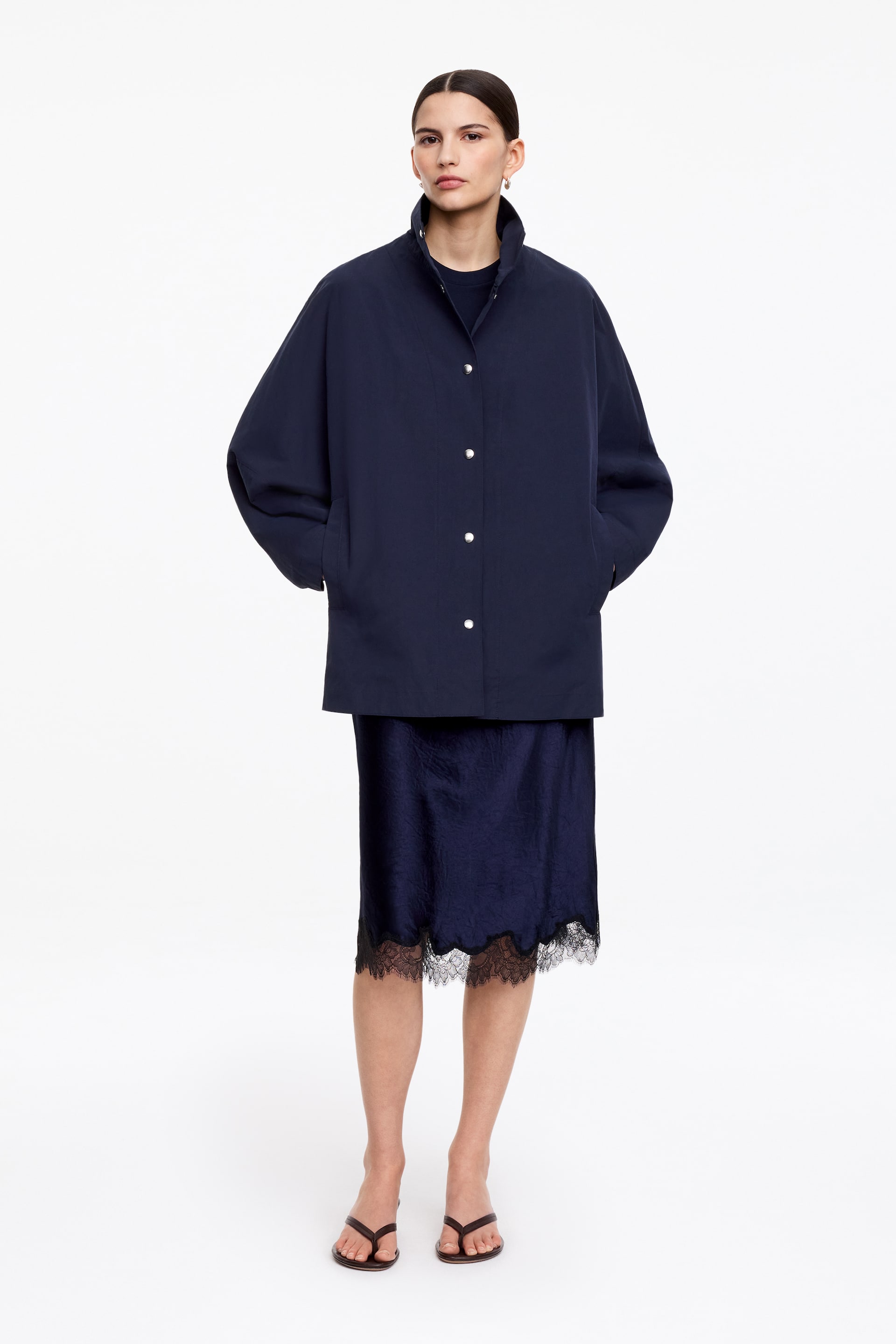 Visa större bild: Den marinblå jackan visas från en framåtvänd vinkel, med en ståkrage och en framknäppning med ljusa knappar. Den har en avslappnad, oversized passform, faller till mitt på låret, och händerna vilar i dess sidofickor.